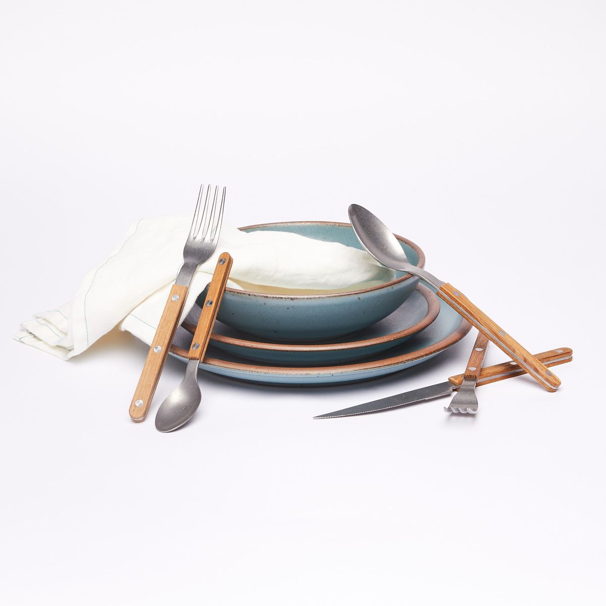 An everyday bowl, side plate, and dinner plate in turquoise stacked with a linen napkin surround by five pieces of teak-handled flatware