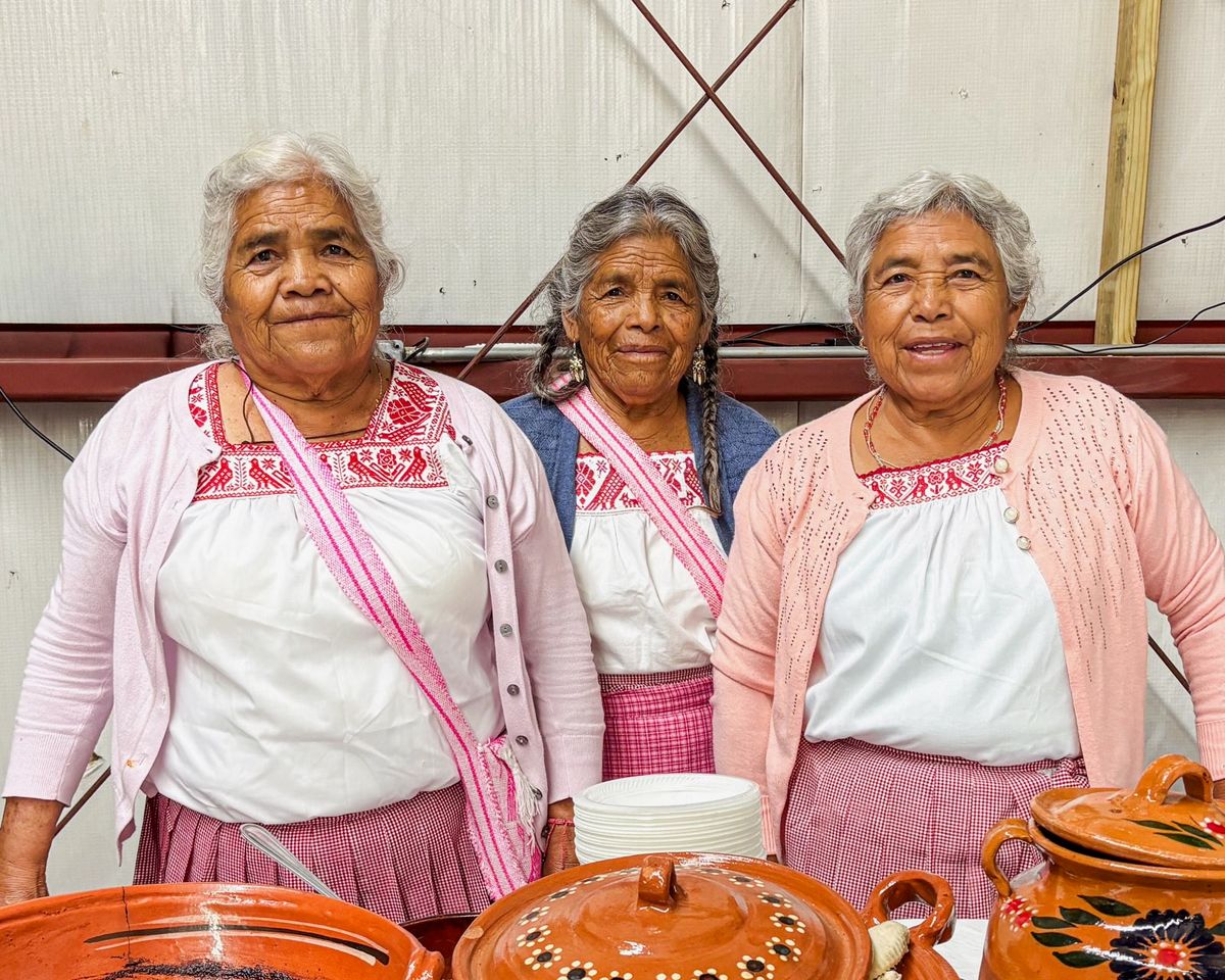 Three people wearing matching white tops with pink embroidery and cardigans standing close together behind a table of large handmade pottery pieces.
