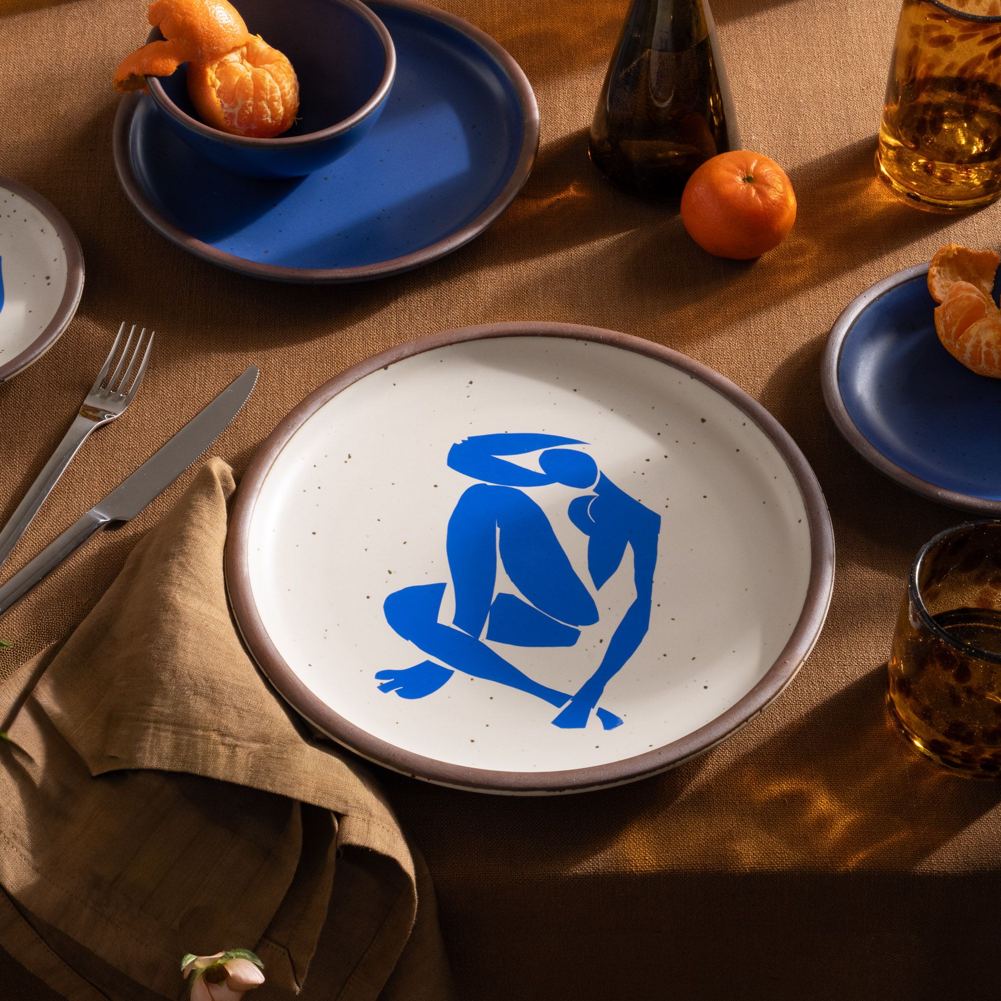 A plate featuring Nu Bleu IV, by Henri Matisse, a bright blue, abstract, silhouetted female figure in a curled, seated position, with sharp cut-out edges, centered on an off-white ceramic plate with an unglazed rim, on a table with blue ceramic dinnerware, clementines, and silverware.