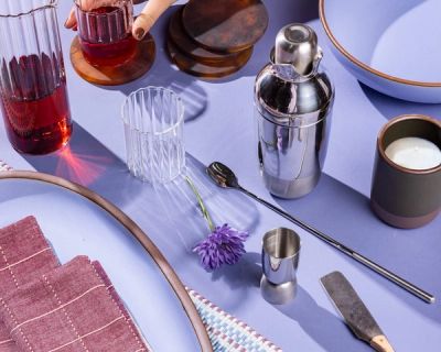 A collection of gifts including a cocktail shaker set, glassware, a mug, and a large oval platter against a periwinkle background.