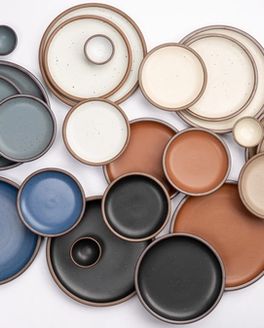 Various sizes of ceramic plates and tiny bowls in a rainbow of earthy colors are artfully laid out together against a white background.