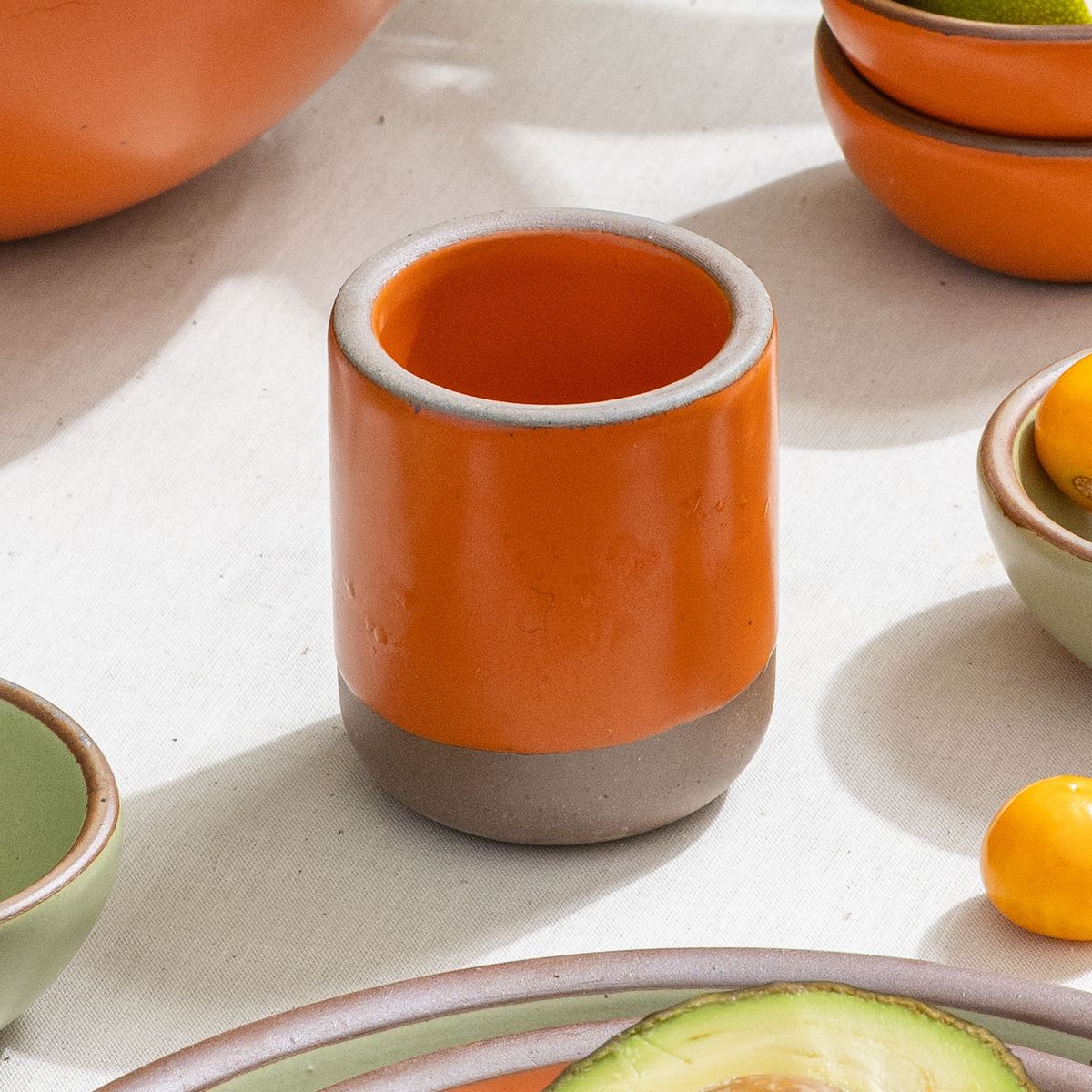 A small, short ceramic mug cup in a bold orange color featuring iron speckles and unglazed rim and bottom base, surrounded by other ceramic pottery in orange and sage green