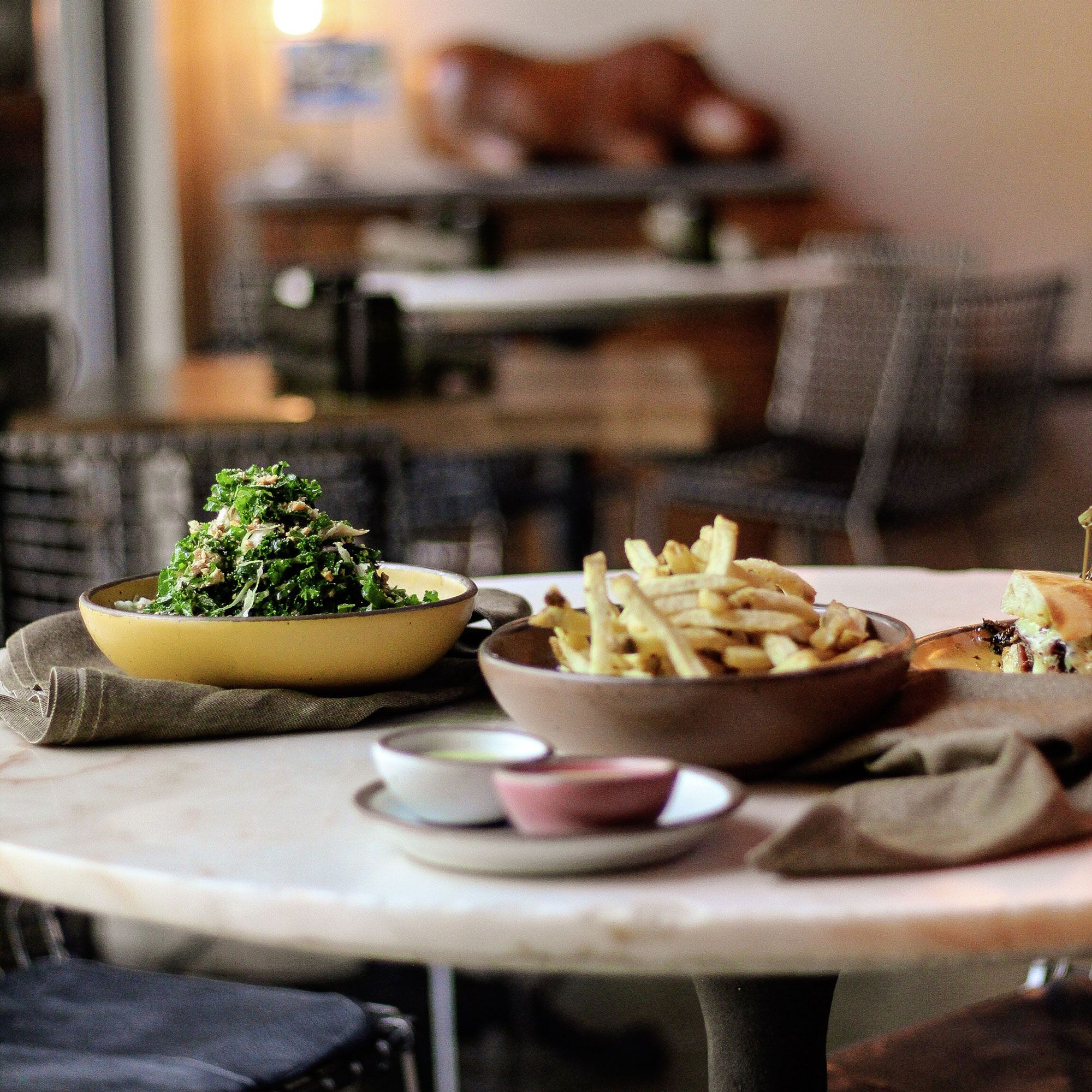 Multiple bowls in a restaurant sit on a table filled with a salad, french fries, a burger, and dipping sauces.