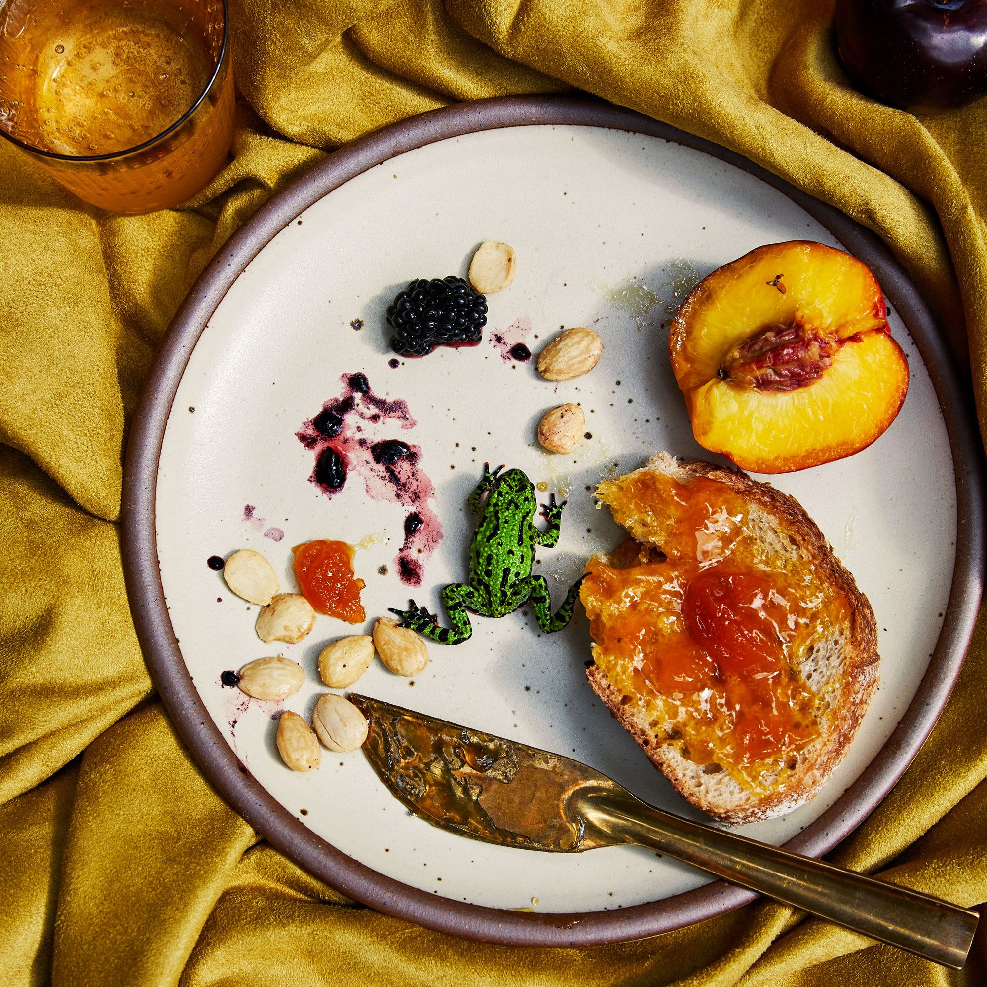 A playful scene happening on a warm white plate - half eaten toast, a peach, a brass knife, and a little green frog