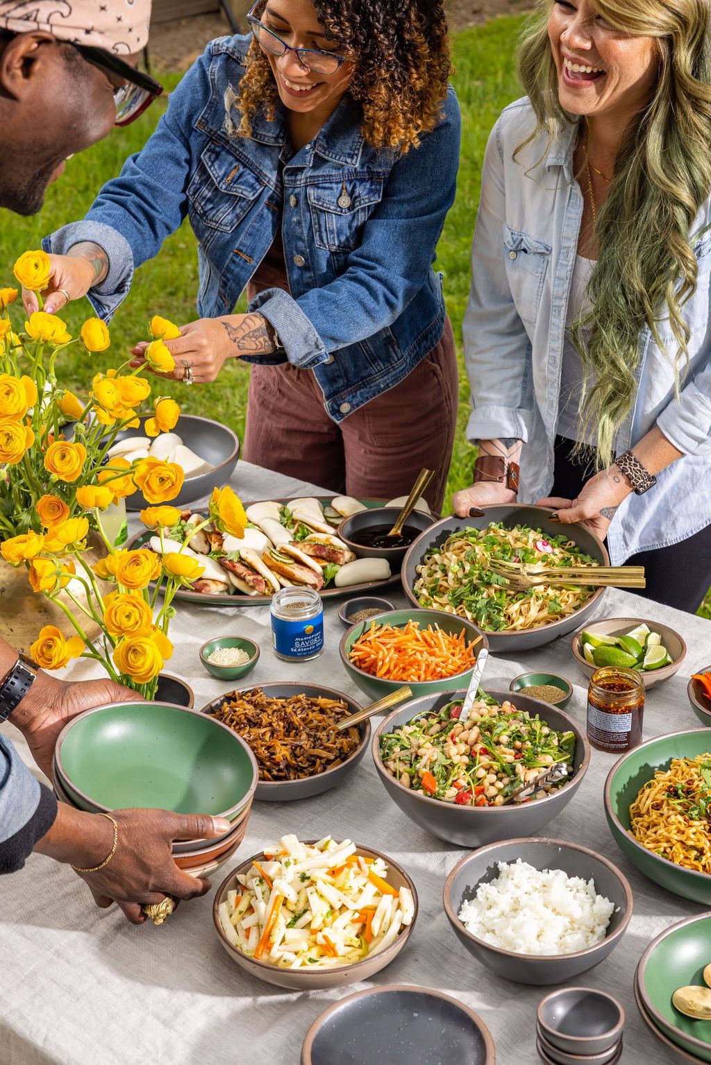 People grabbing food from an abundant table outside filled with bowls of rice, noodles, meats, and veggies.