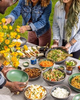 People grabbing food from an abundant table outside filled with bowls of rice, noodles, meats, and veggies.