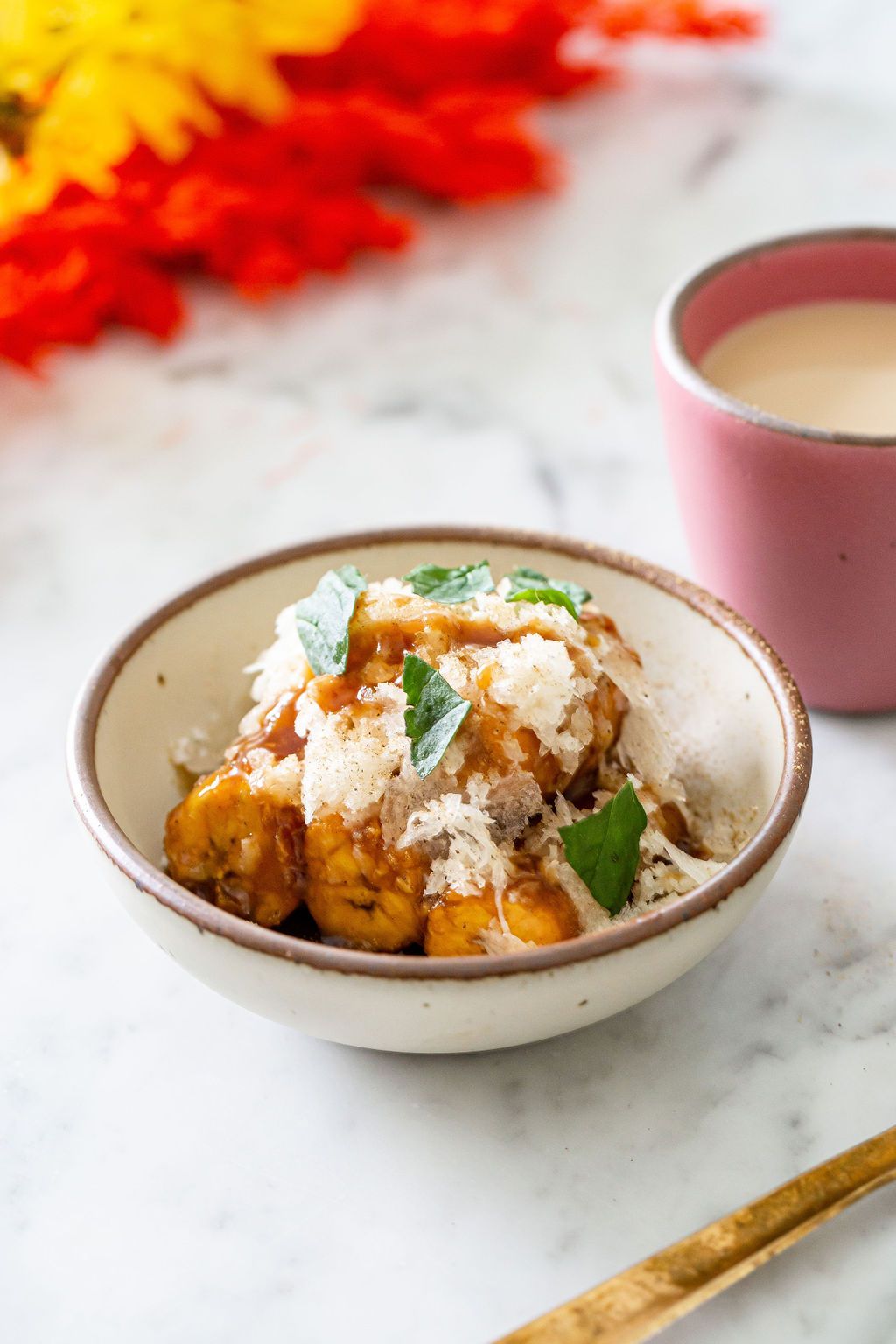 On a table sits a beautiful dish of caramelized plantains in a ceramic off-white bowl, with a pink ceramic cup of milk behind it.