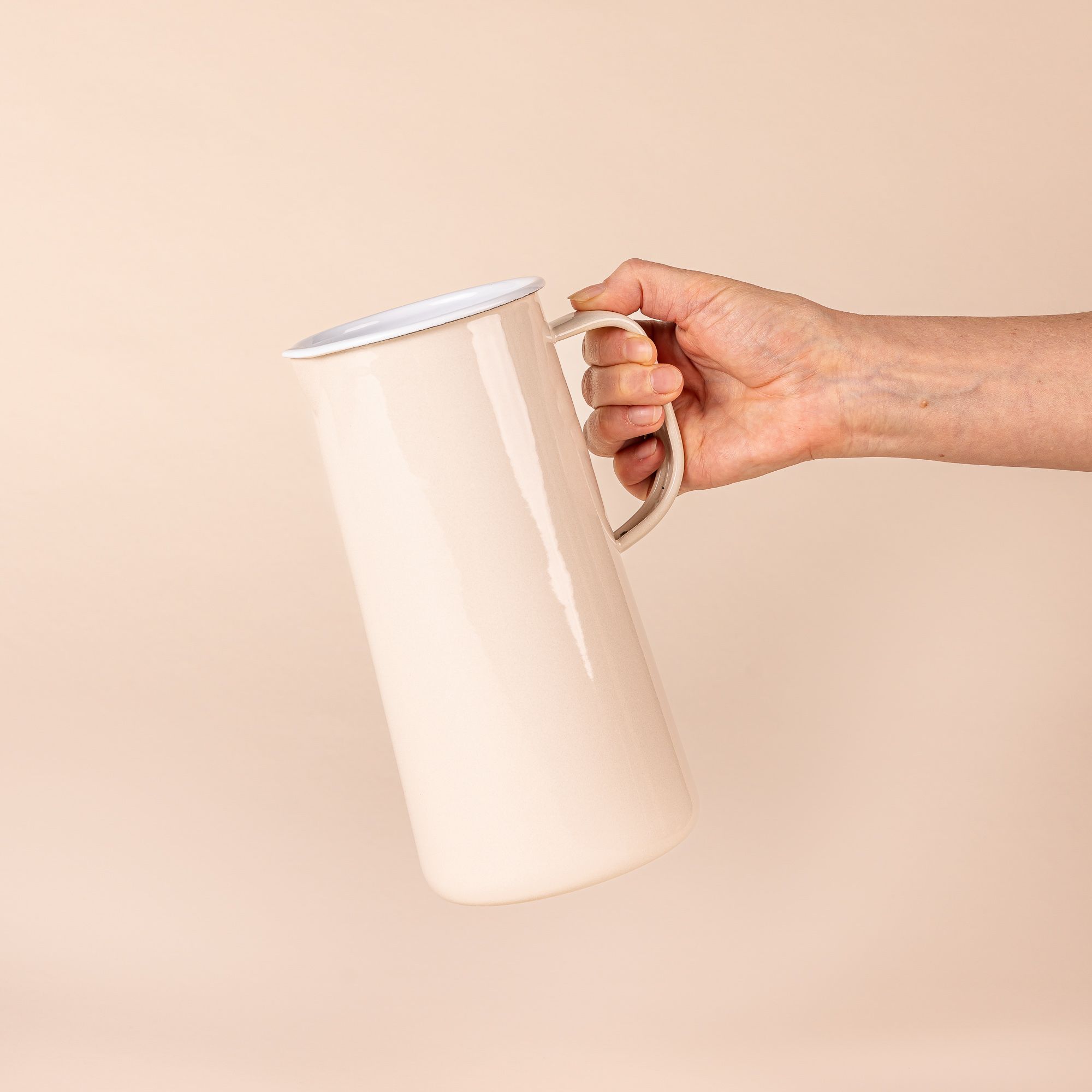 A hand holding a shiny cream cylindrical jug, with white interior, a spout on the left, and a handle on the right