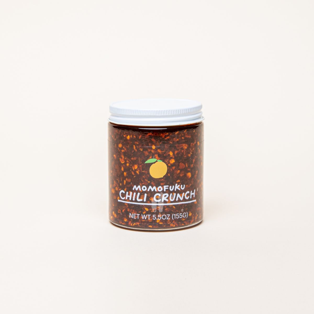 A clear jar with a white cap filled with a warm brown sauce with chili flakes- the jar has a label that reads, "Momofuku Chili Crunch"