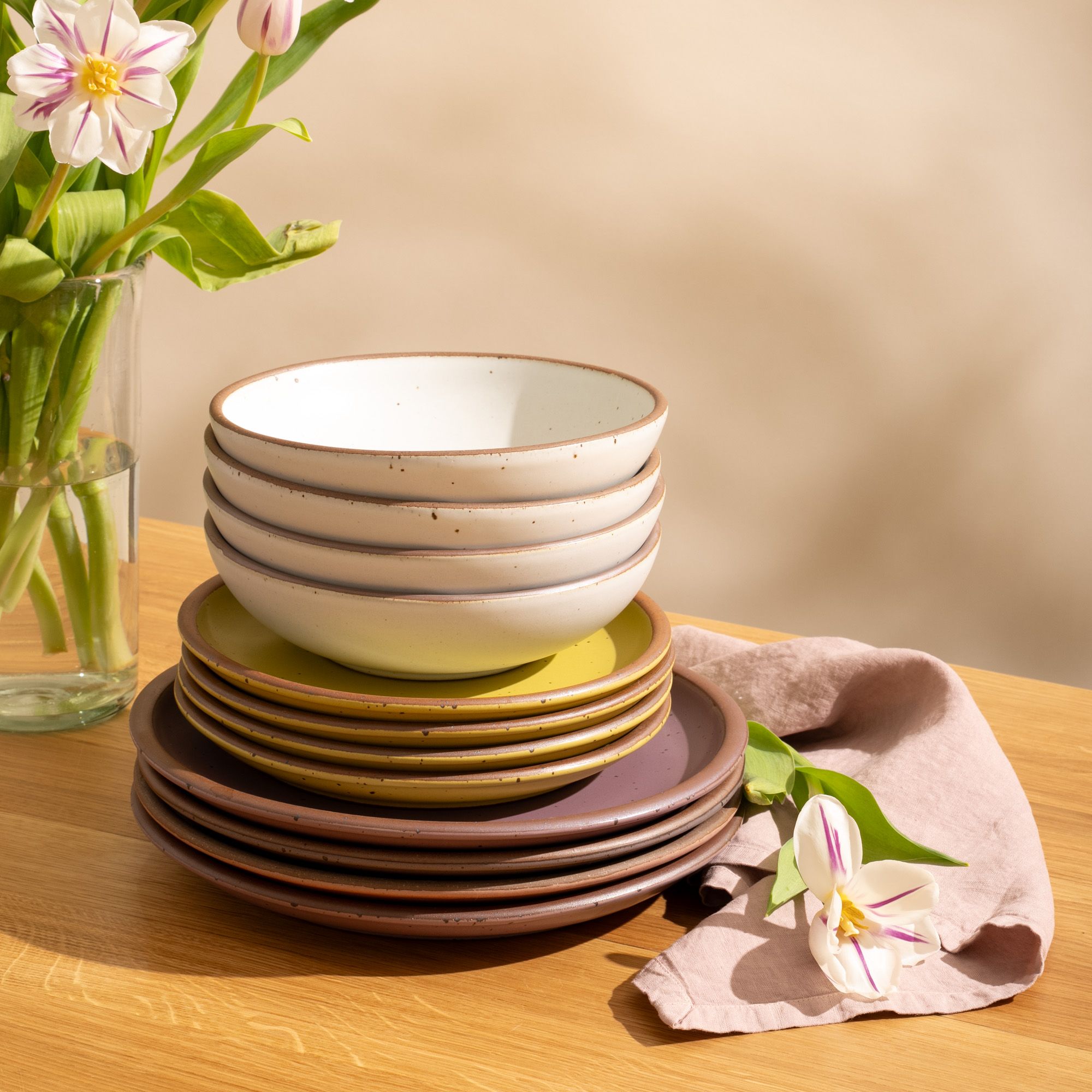 4 ceramic dinner plates in soft mauve purple, 4 side plates in cool white, and 4 shallow dinner bowls in zesty yellow with iron speckles and unglazed rims, stacked together on a table with a flower and pale pink napkin.