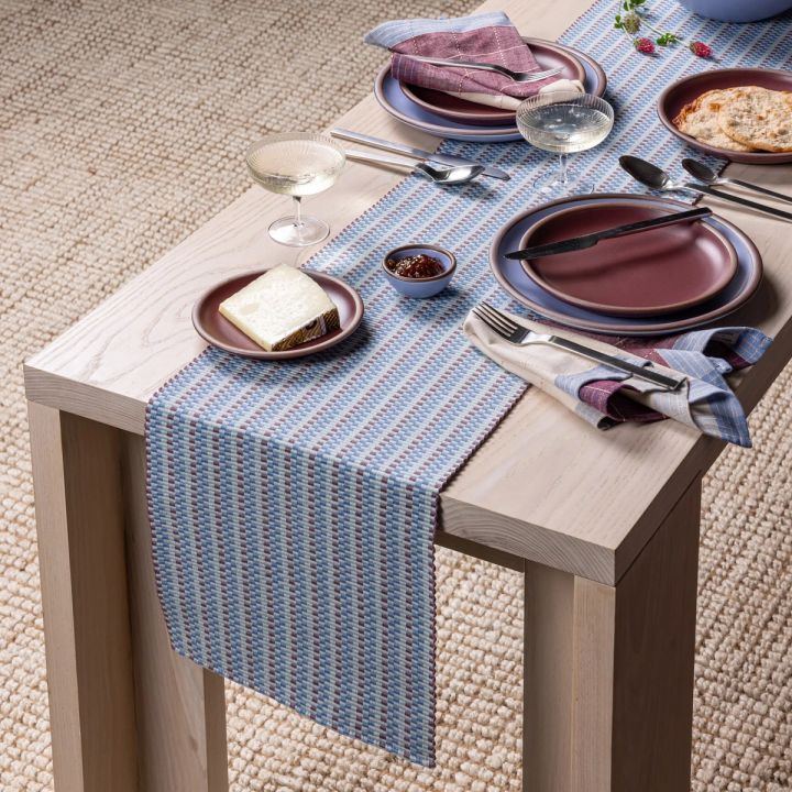 A striped woven runner in soft blue tones lays on a table with dishes, coupe glasses, napkins, and silverware.