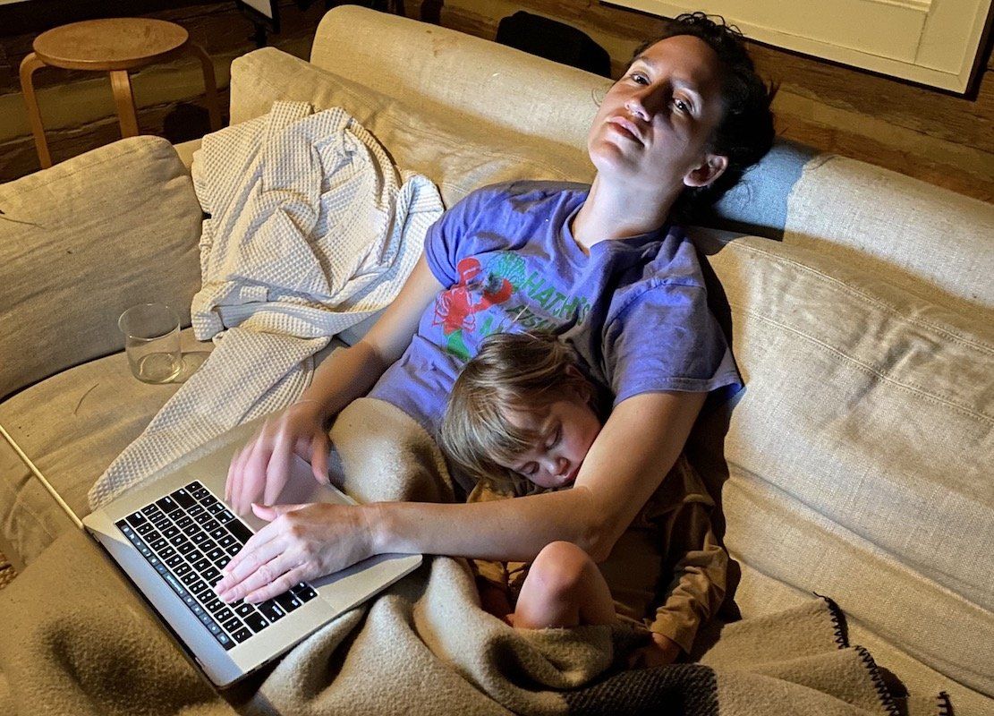 Connie sits on a cream couch with a blanket, her laptop open on her lap, and her child curled up next to her.