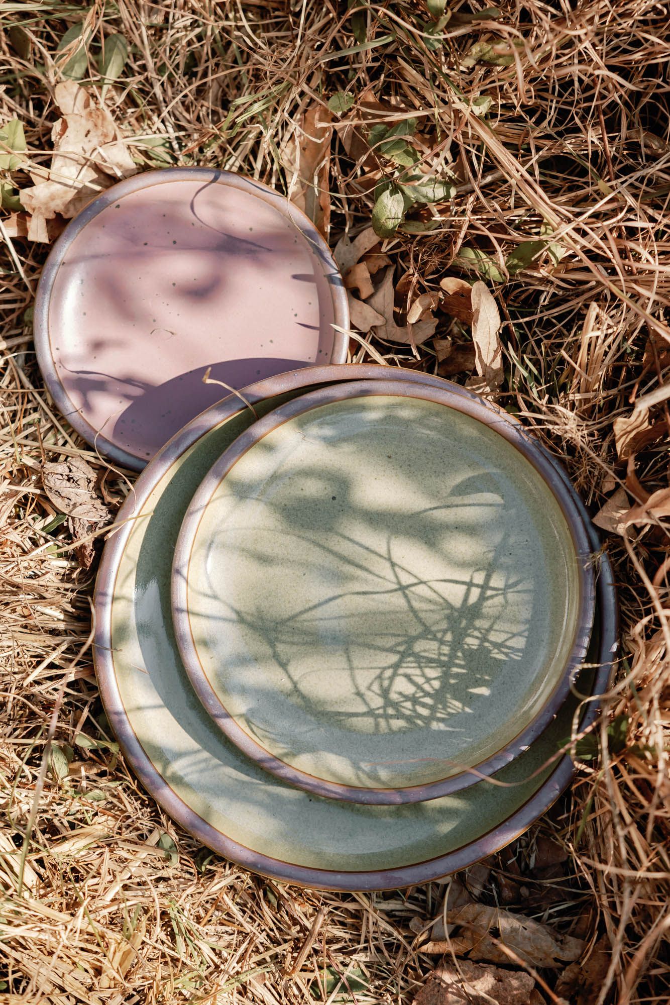 3 Plates of varying sizes in reflective dappled grey-green and dusty mauve pink artfully arranged in dried grass with shadows.