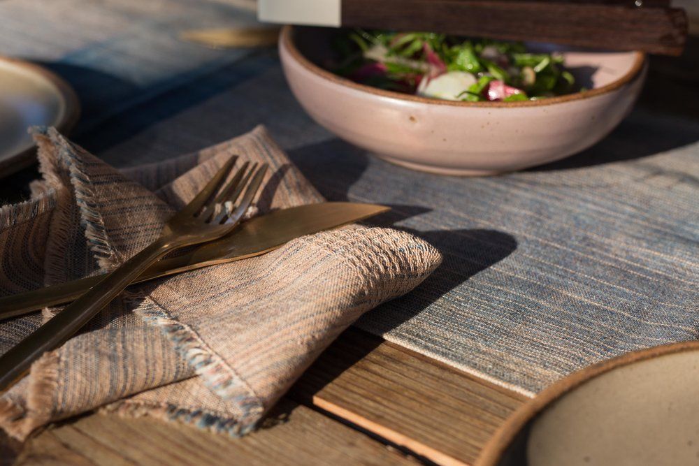White and blue woven napkins sit on a table with shiny flatware, and a ceramic bowl.