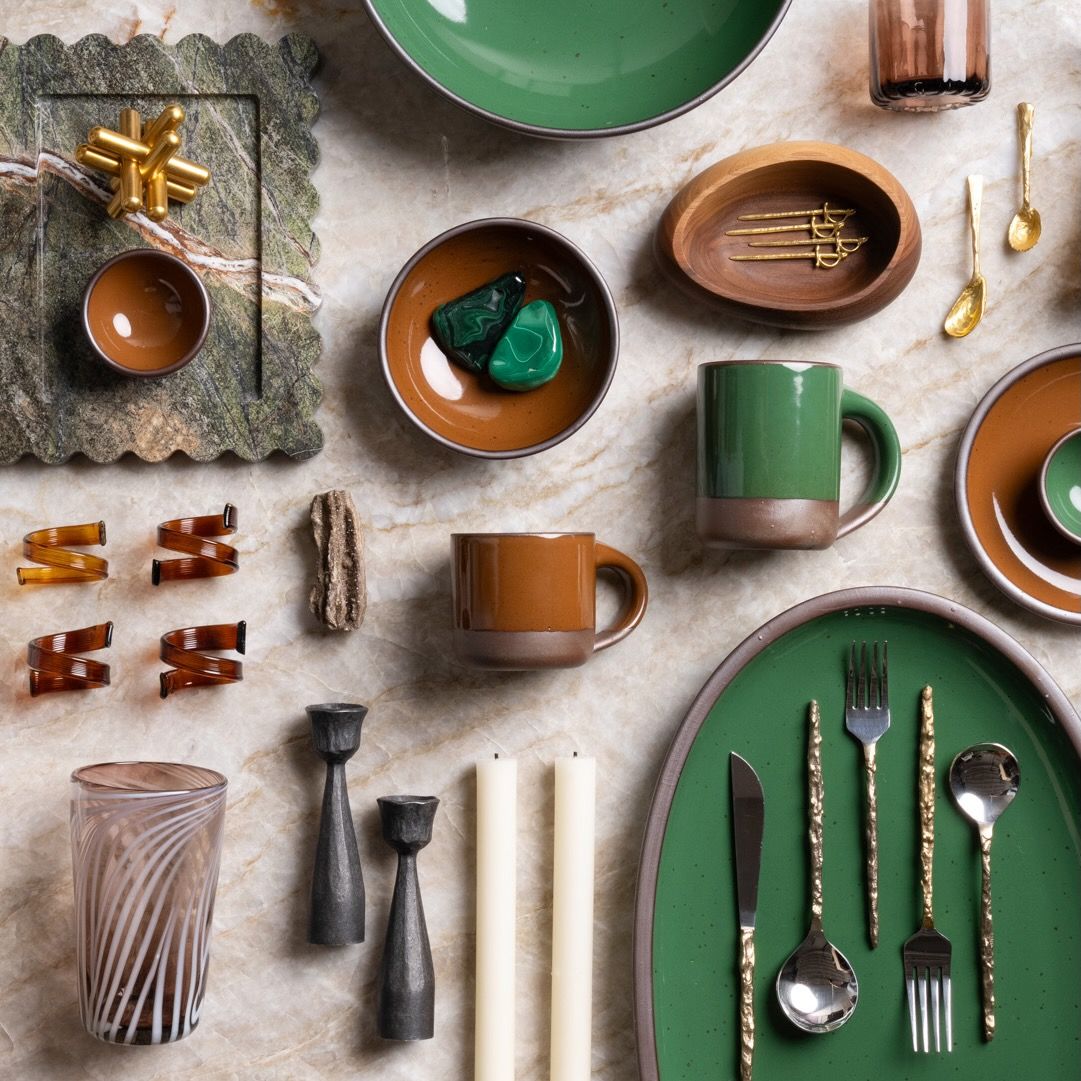 A styled flat lay of East Fork pottery, candles, glassware, and gold-toned flatware arranged artfully on a marble surface.