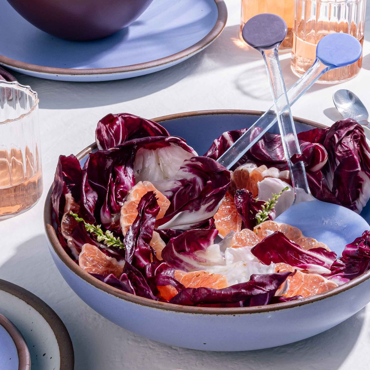 A large shallow serving ceramic bowl in a periwinkle color filled with salad and colorful serving spoons