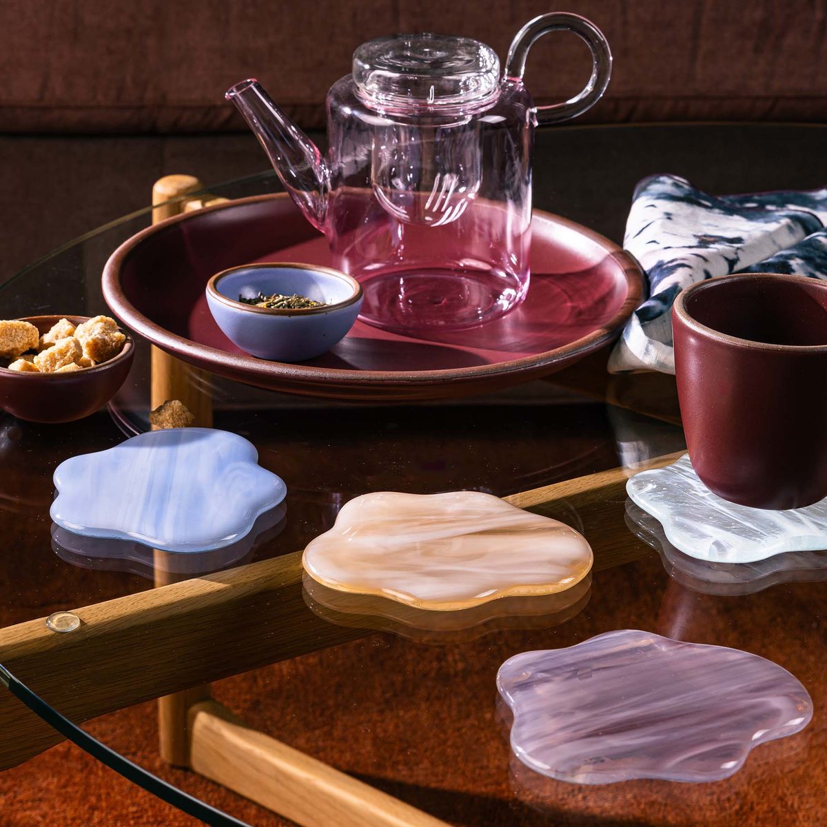 On a table sits 4 glass coasters shaped like clouds with a small cup, glass teapot, and tea fixings.