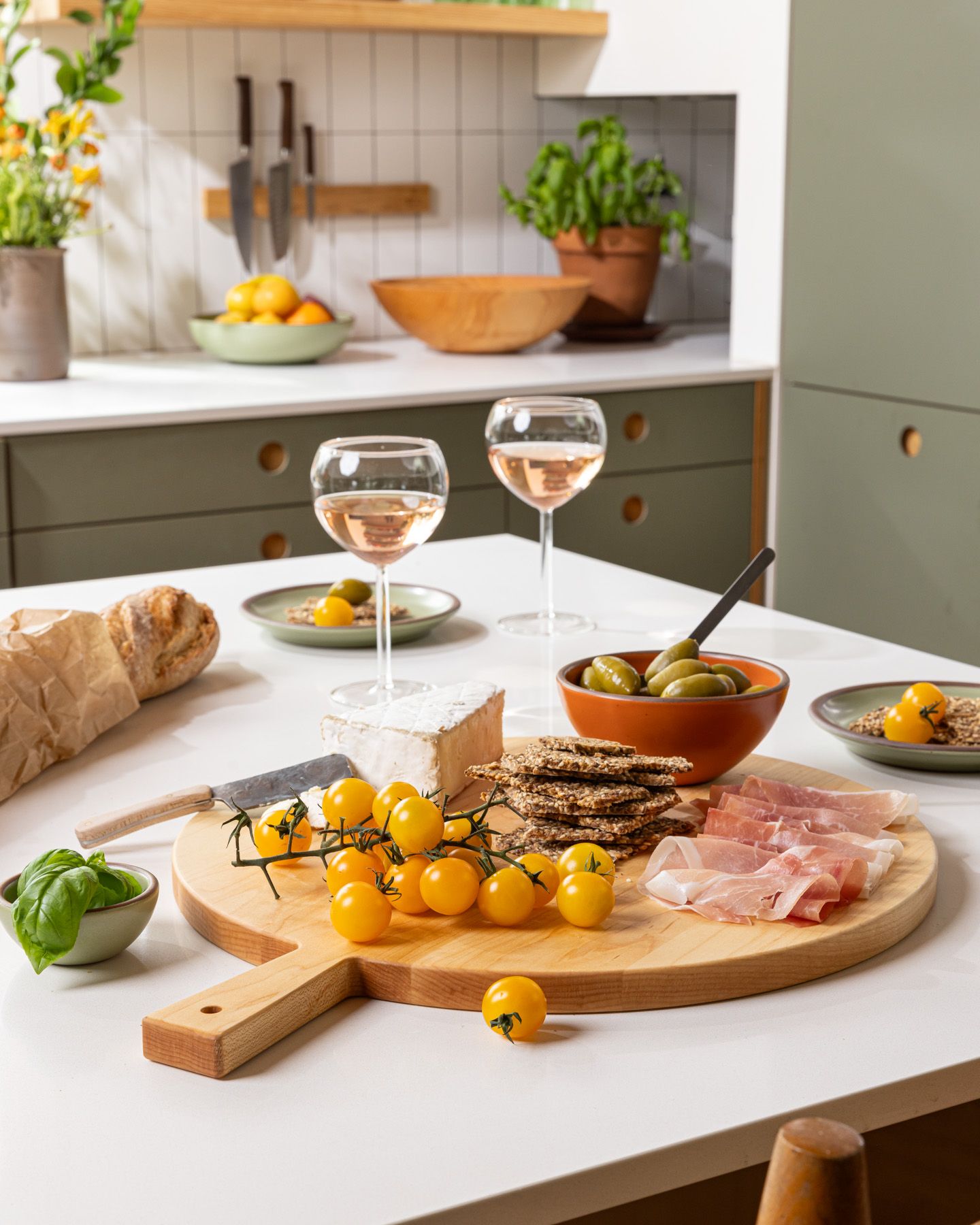 On a kitchen island is a round wooden serving board filled with charcuterie and surrounded by small plate, bowls, and wine glasses