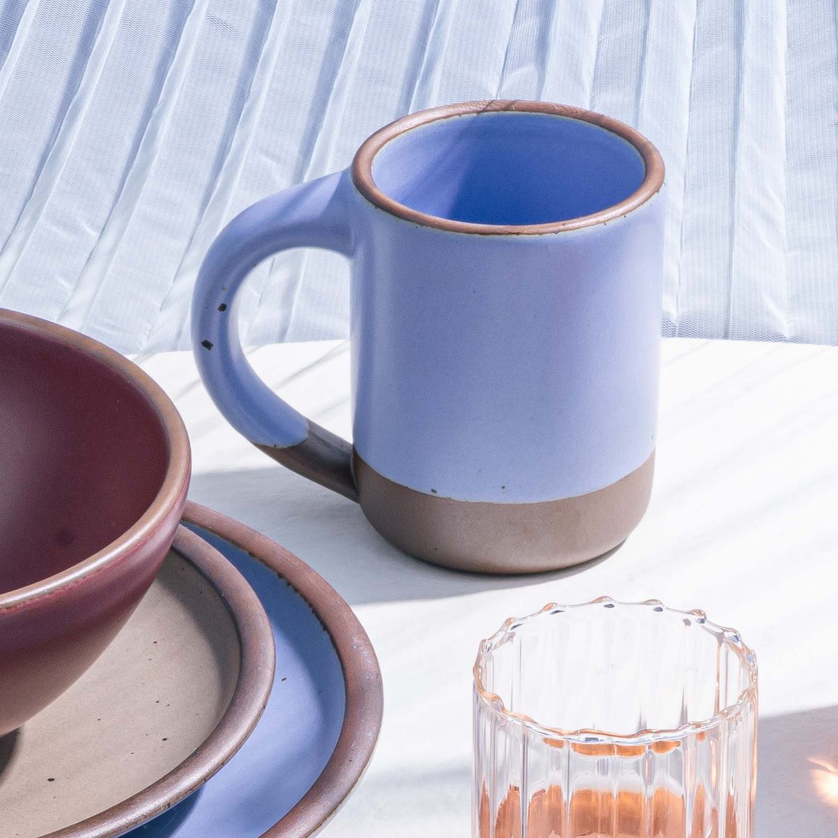 A medium sized ceramic mug with handle in a periwinkle color on a table with a glass and plates