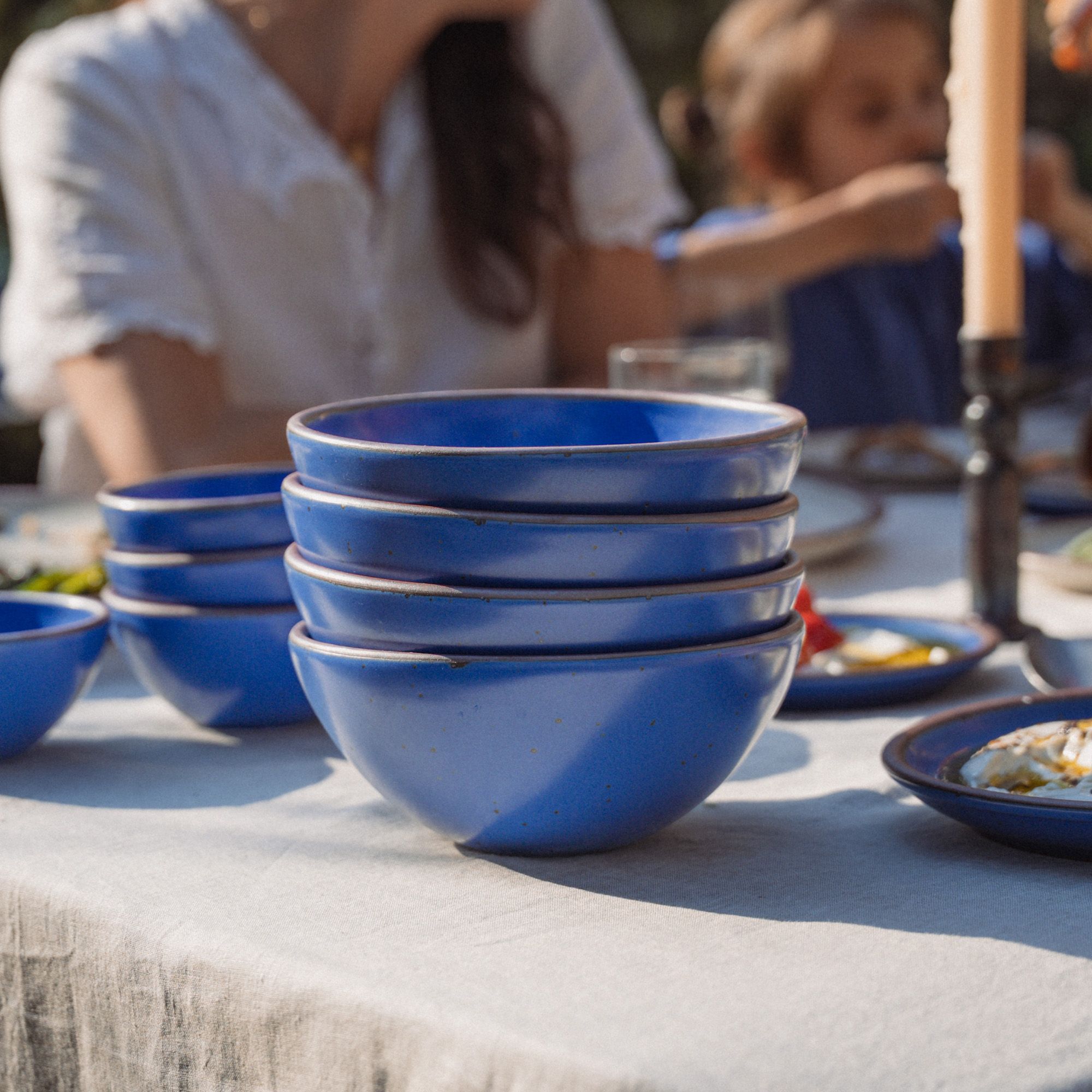 Outside on a table is a stack of 4 medium rounded ceramic bowls in a cobalt blue color featuring iron speckles and an unglazed rim, along with more bowls, foods, and a candle.