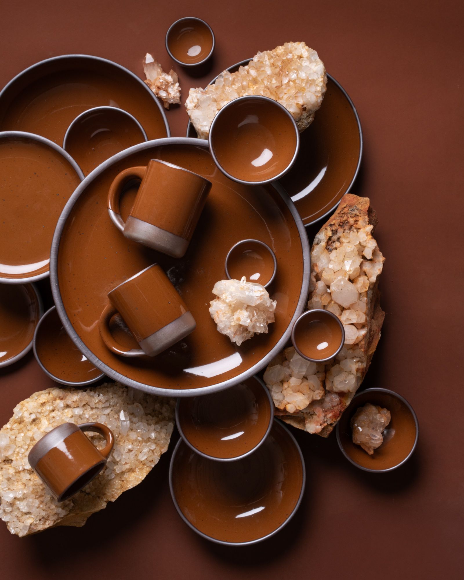 Glossy brown ceramic mugs and bowls arranged with plates and large quartz crystal clusters on a brown background.
