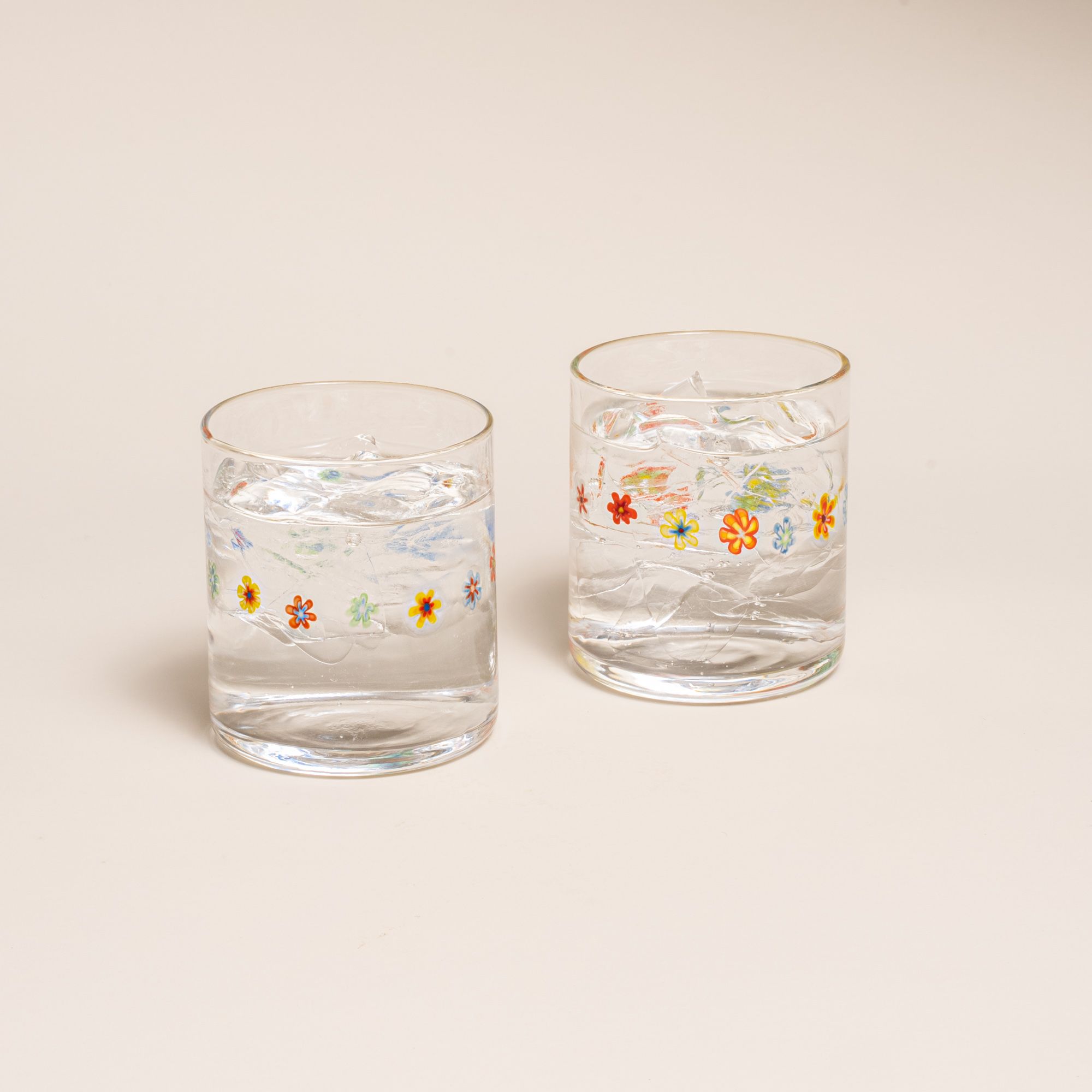 Two clear glasses with a ring of multicolored little flowers, filled with water, on a cream background.