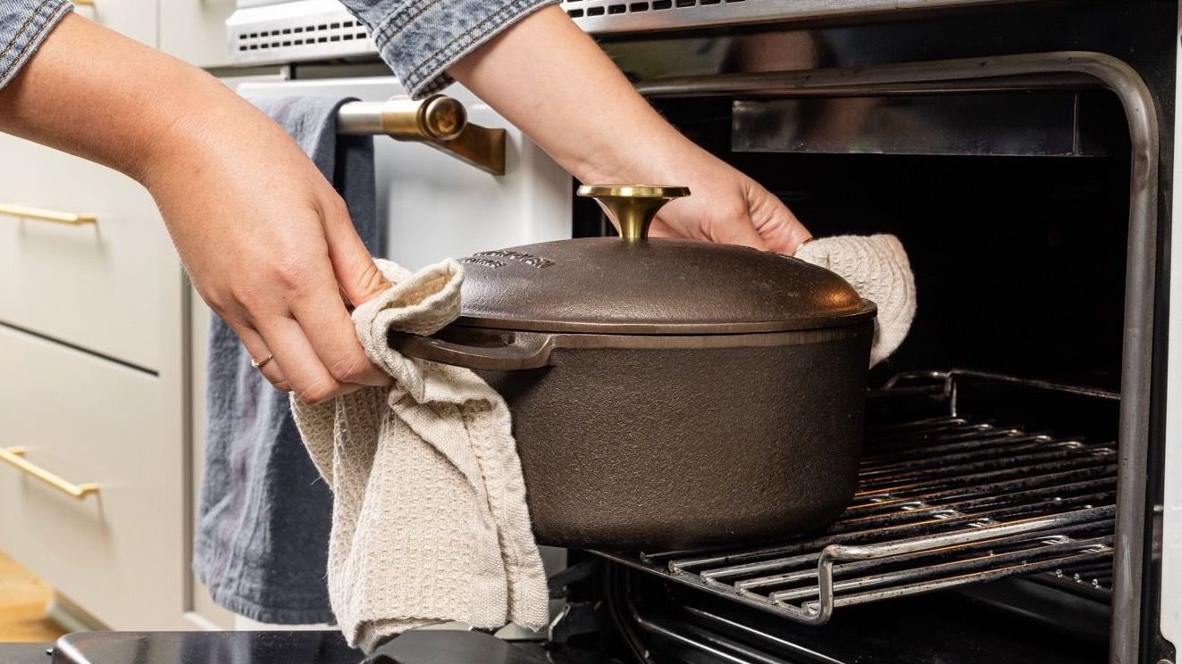 Hands hold a kitchen towel to carry a cast iron dutch oven with a handle on each side and cast iron lid out of an oven.