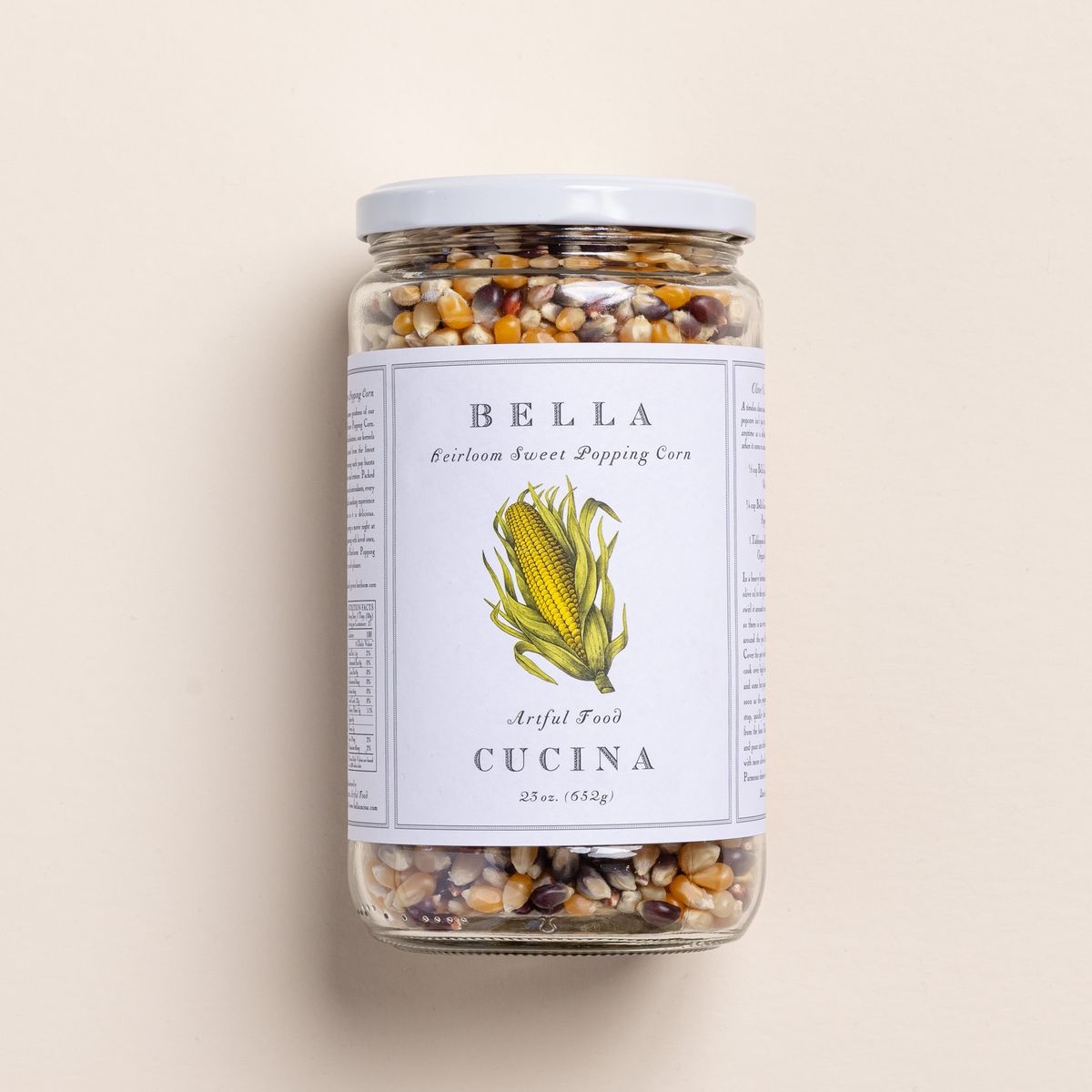 A jar with a white lid filled with popcorn kernels, and a large label with an illustration of corn and reads 'Bella Cucina Heirloom Sweet Popping Corn.'