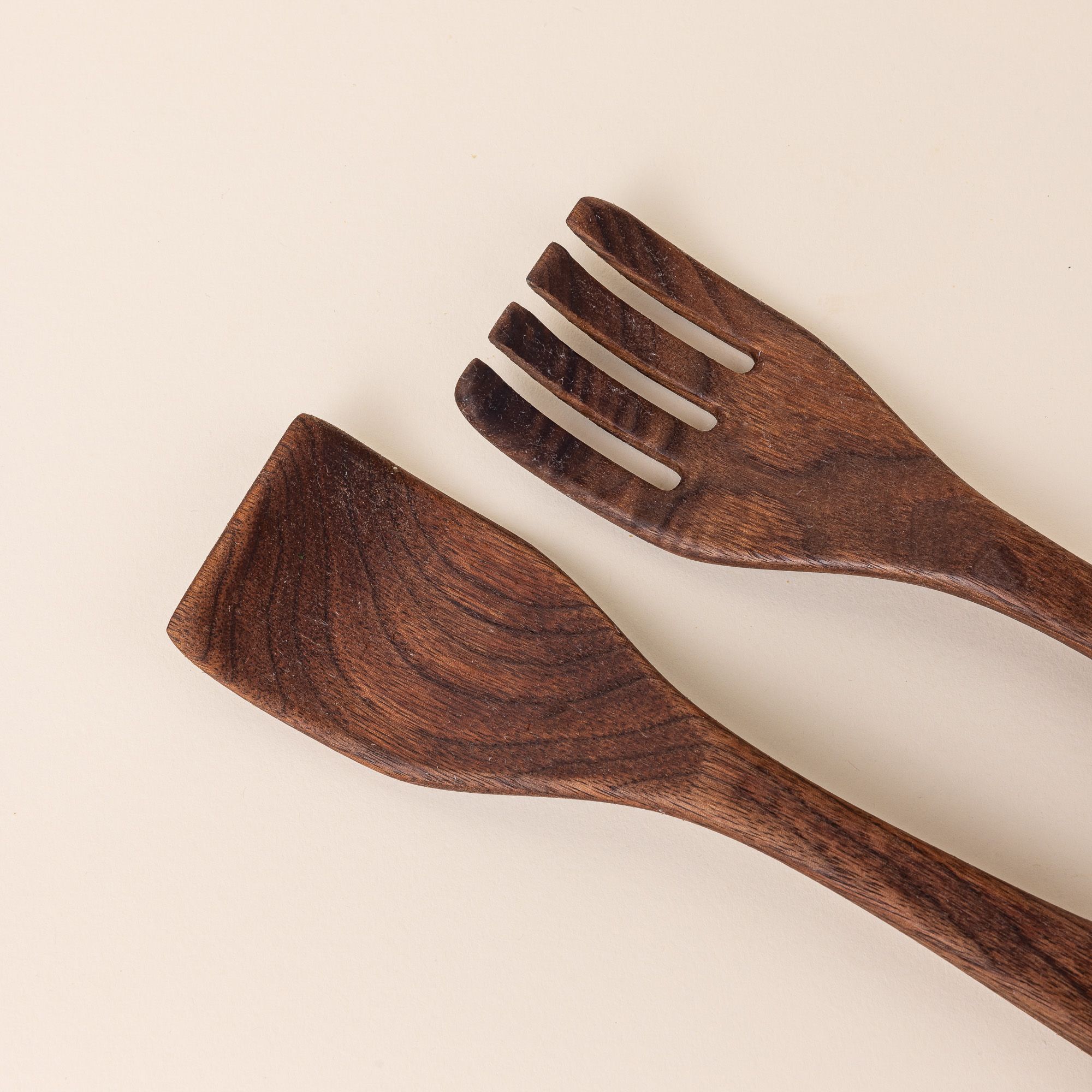 Closer look at the end of two long walnut wood serving utensils. The left utensil has a shape like a spatula, the right has a shape like a fork.