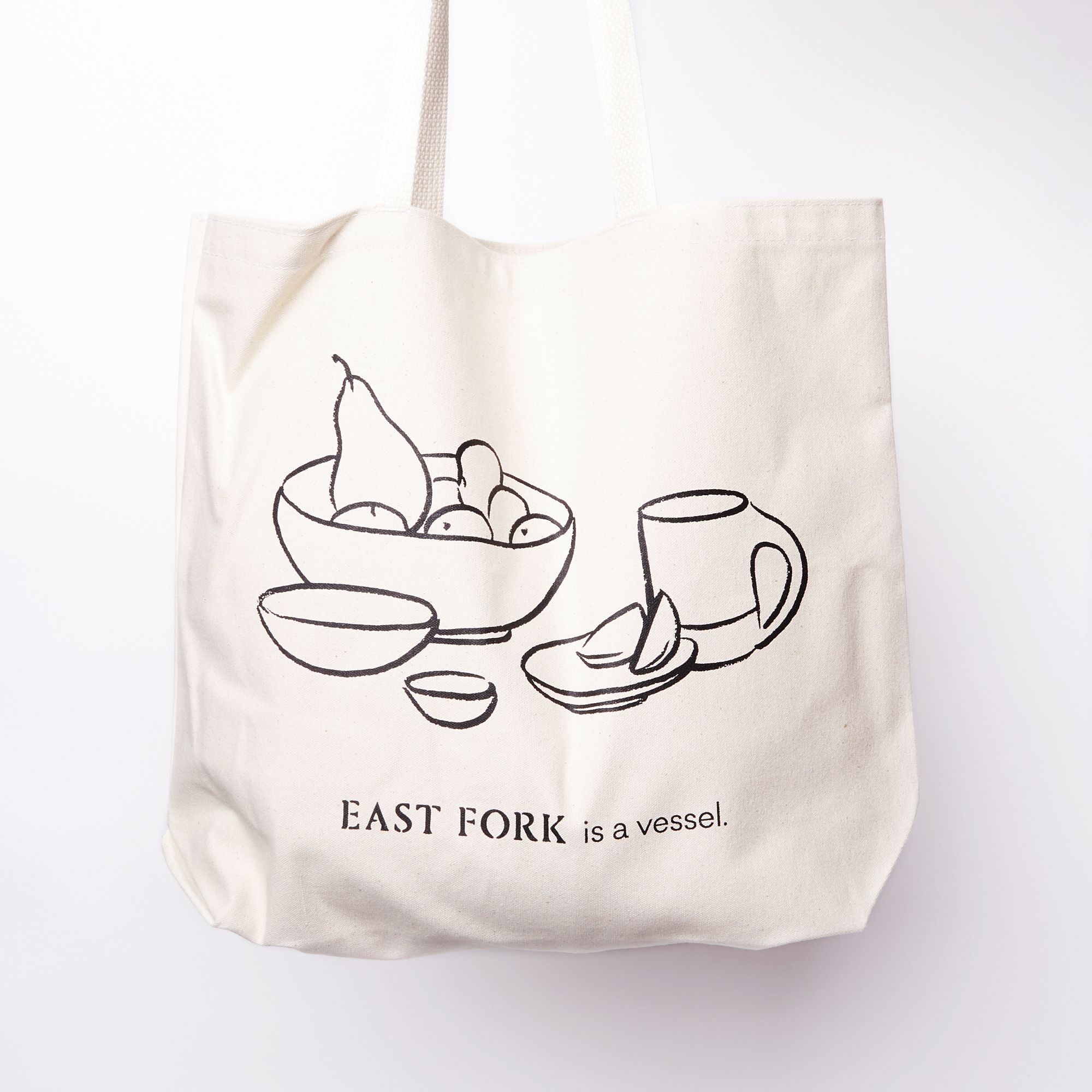 Cream canvas tote with a charcoal illustration on the front of a bowl with fruit, little bowls, plate, and a mug. Underneath, it reads 'East Fork is a vessel'.