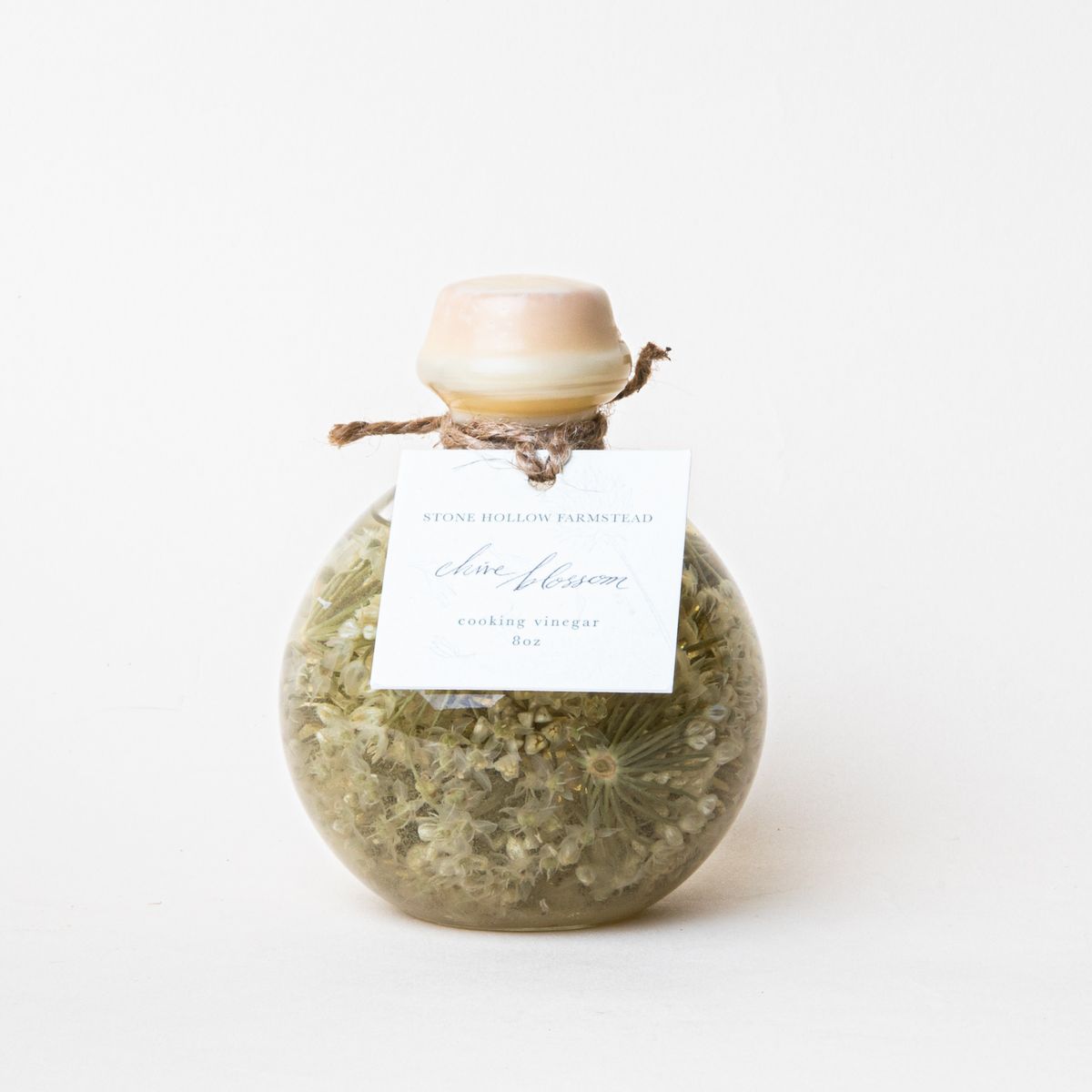 Round jar filled with flowery liquid in a sage green color with a wooden cap and a small white label