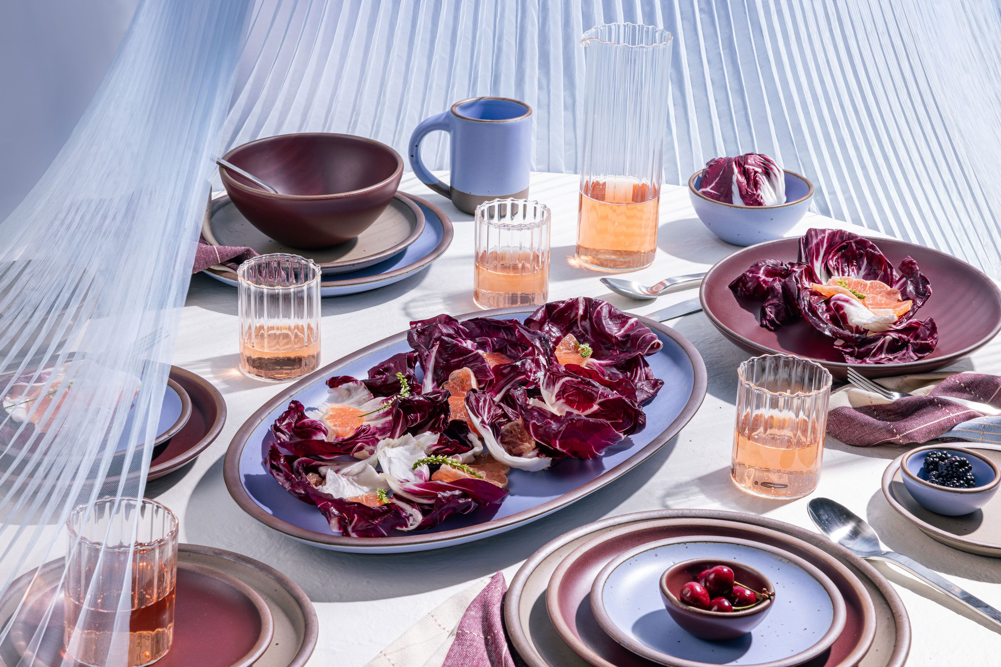 A table filled with ceramic pottery in periwinkle and plum colors, a salad, and glasses filled with orange liquid