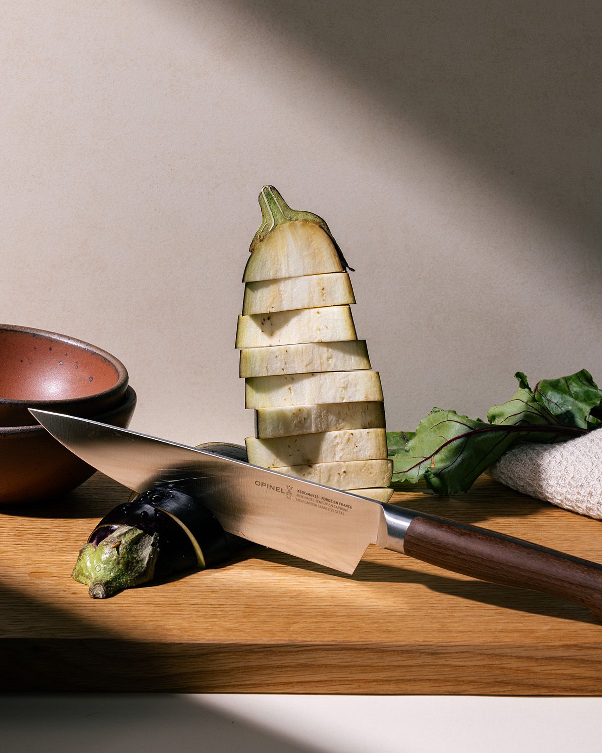 A chef's knife is propped up on a cutting board with slices of eggplant next to the blade and towered behind. The knife has a sharp steel blade with a beveled dark wood handle