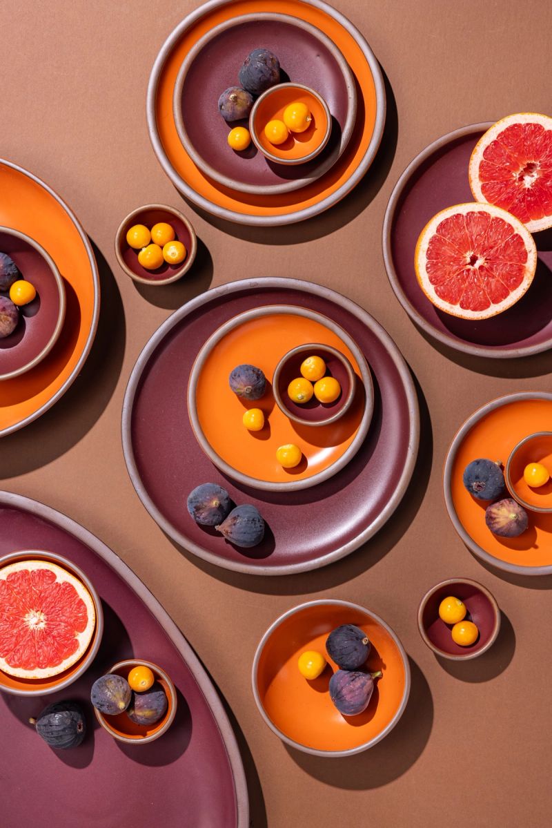 Artistically styled ceramic plates on a light brown background in colors of deep plum and bright orange, holding cut grapefruits, figs and tomatoes. 