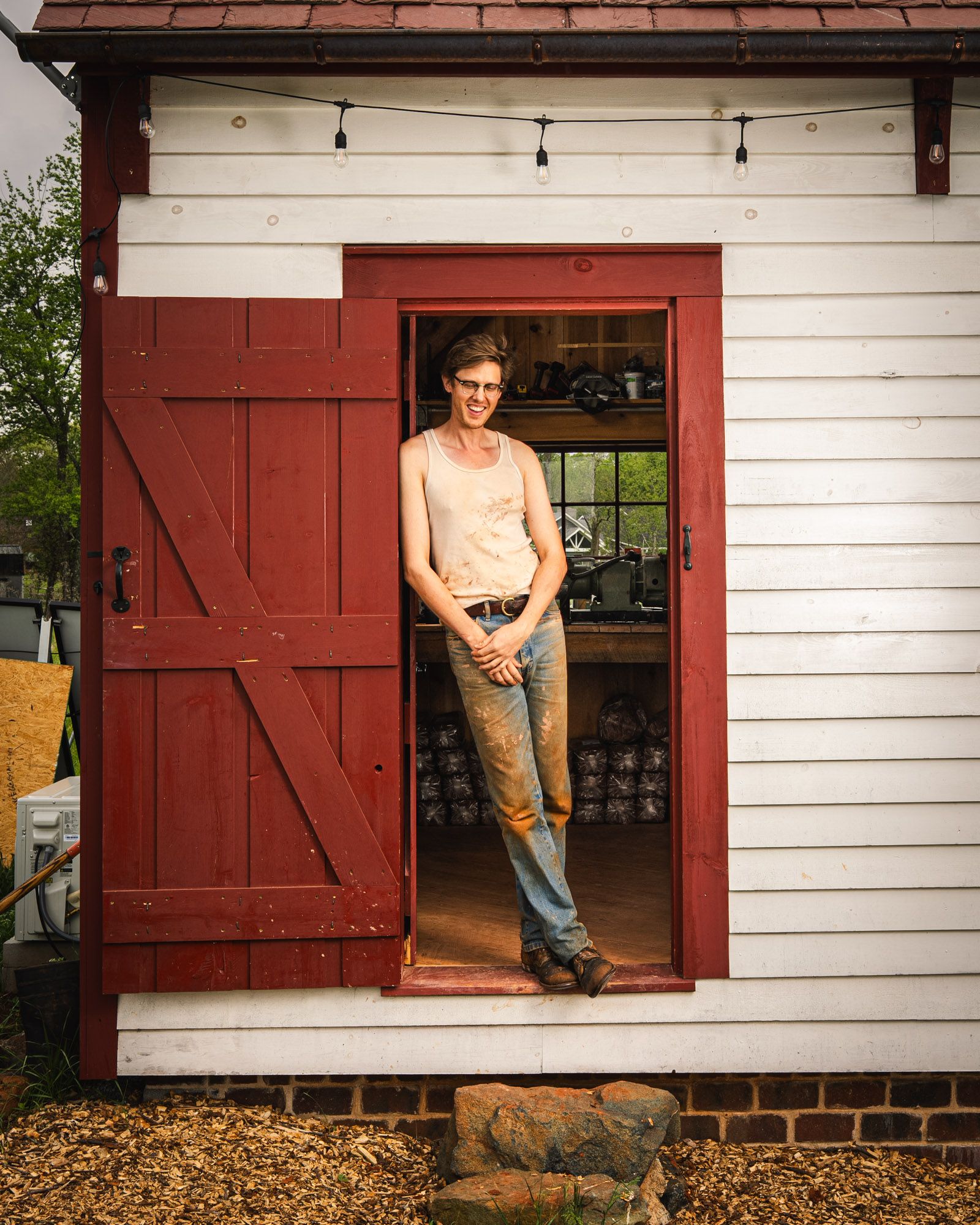 John standing and leaning on the doorway and smiling at his small white cabin pottery studio.