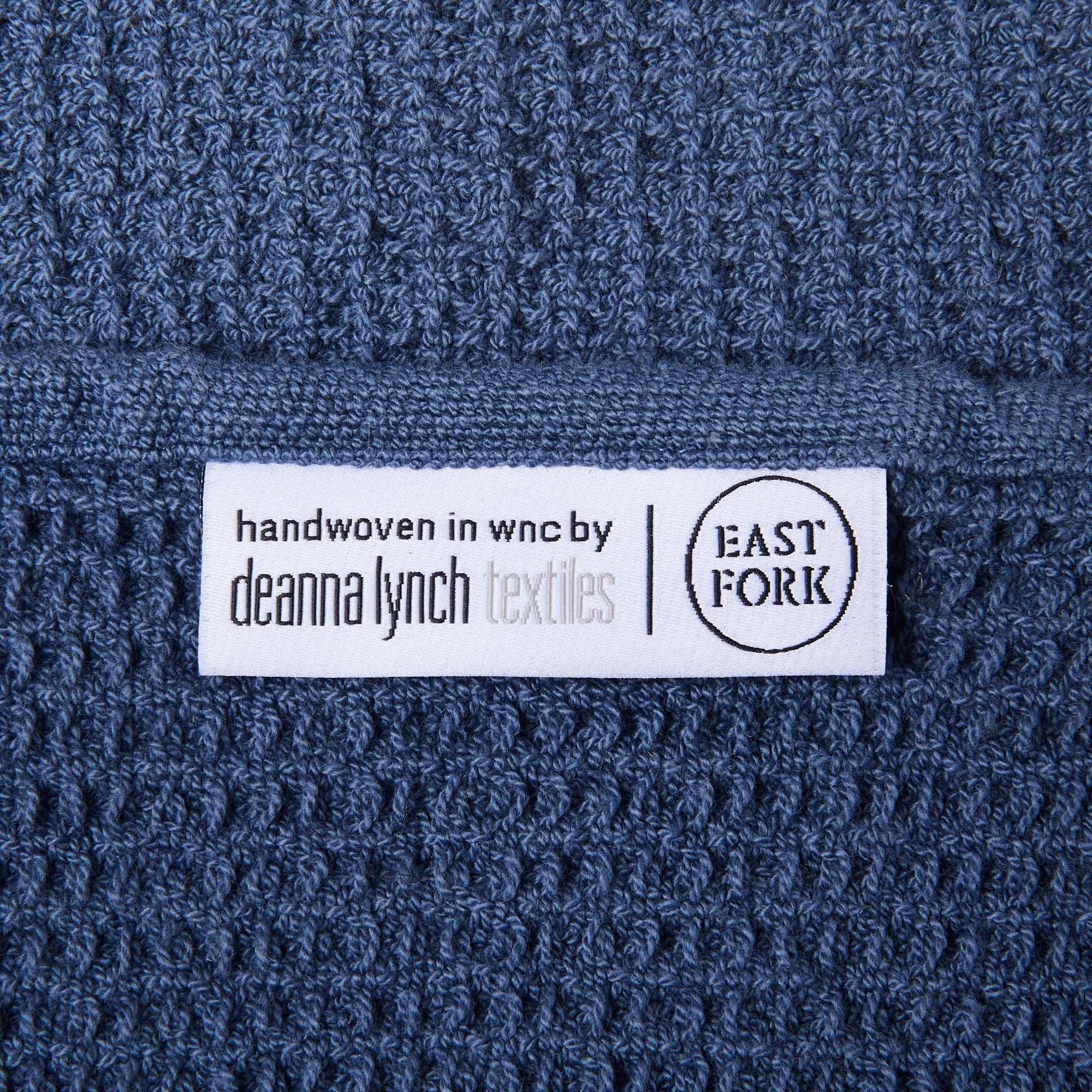 Close up of a blue waffle towel with a white label that reads "Handwoven in WNC by Deanna Lynch Textiles" next to the East Fork logo