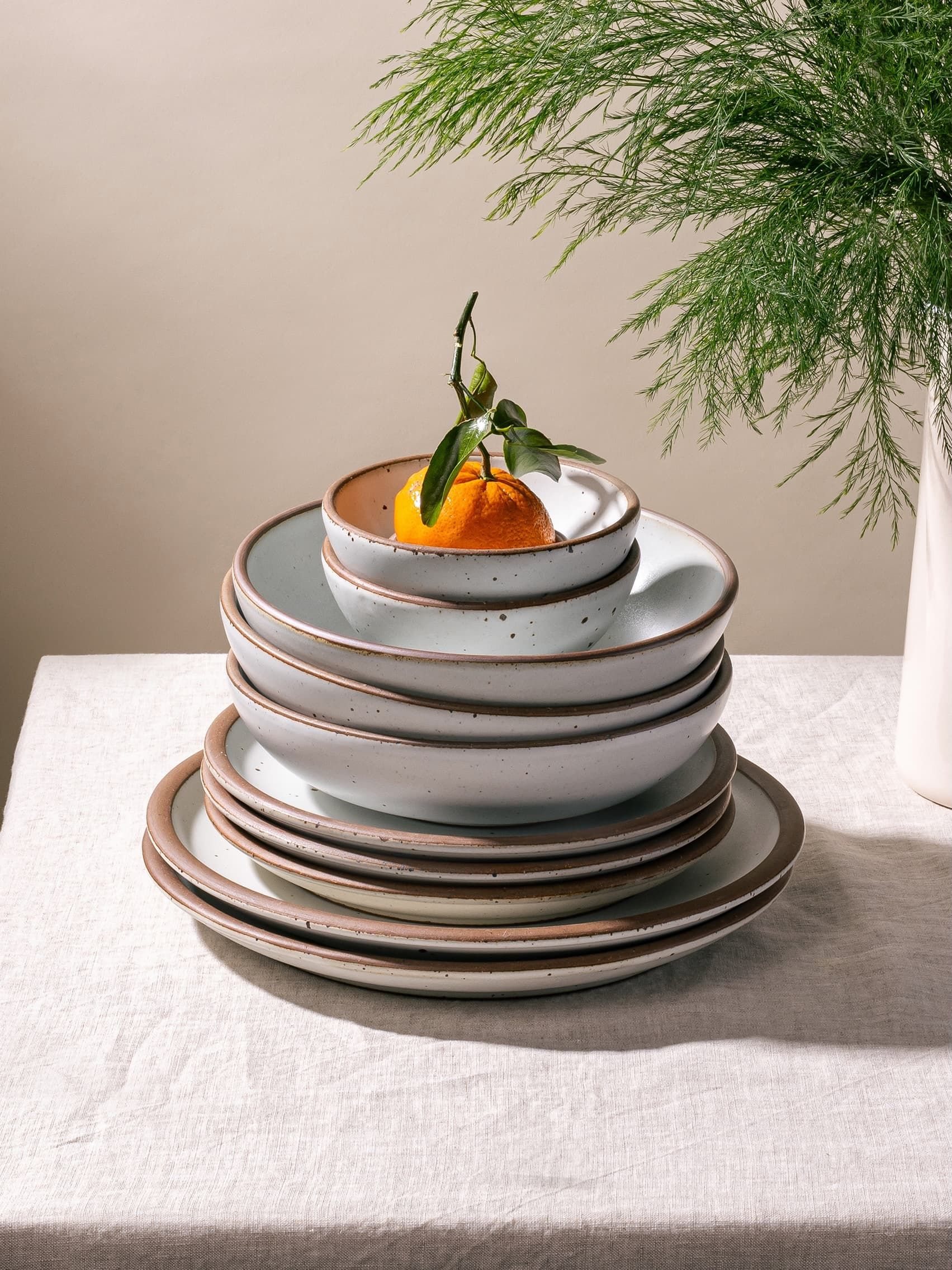 On a table with a linen tablecloth, there is a large stack of ceramic plates and shallow bowls, all in off-white or cool white colors, with an orange sitting on top. A cream pitcher with a fern sits to the right.