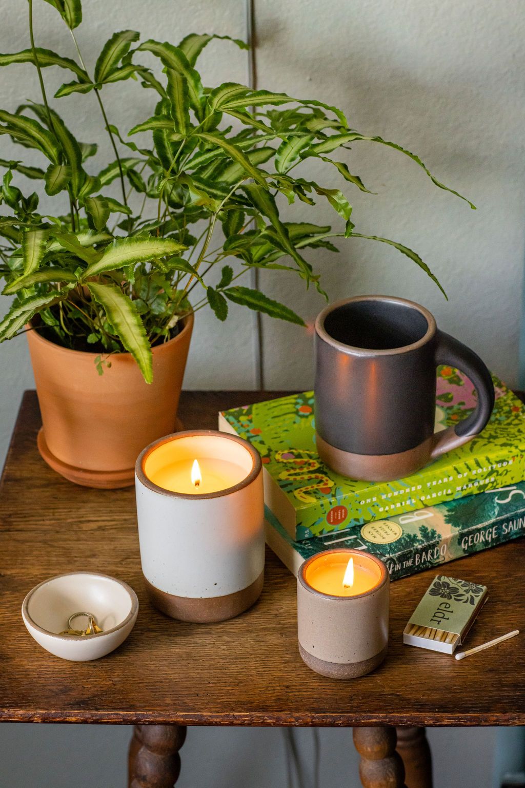 On a bedside table, candles in ceramic vessels are lit, there's also a stack of books with the East Fork Mug in Black Mountain on top.