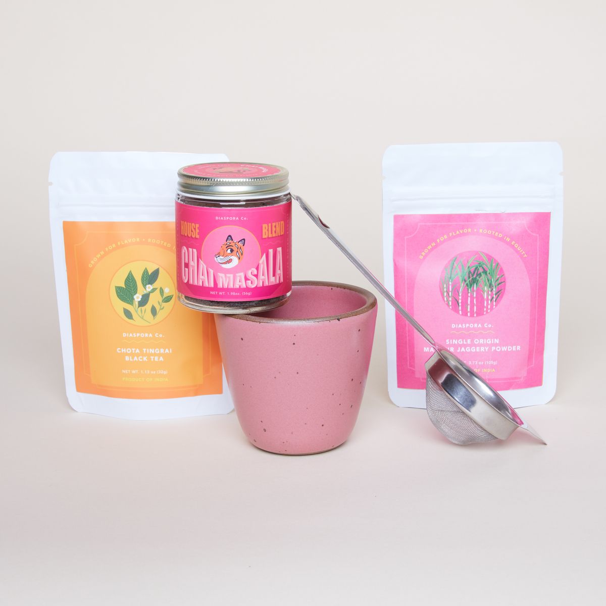 Two Chai Kuhlads – handleless mugs – in Rococo, a hot pink seasonal glaze made by East Fork Pottery. Photographed with two packages of chai mix. Photographed with a stainless tea strainer, a small glass container with a hot pink label holding Diaspora Co.'s house chai blend and two white plastic bags of more tea from Diaspora. Each have a bright label, in orange and pink.