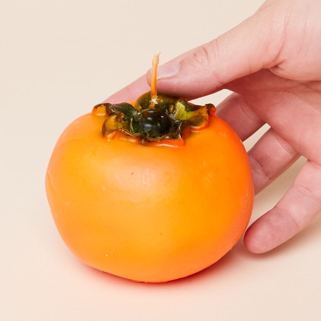 A hand touches a round orange candle with green texture at the top, to suggest a whole persimmon