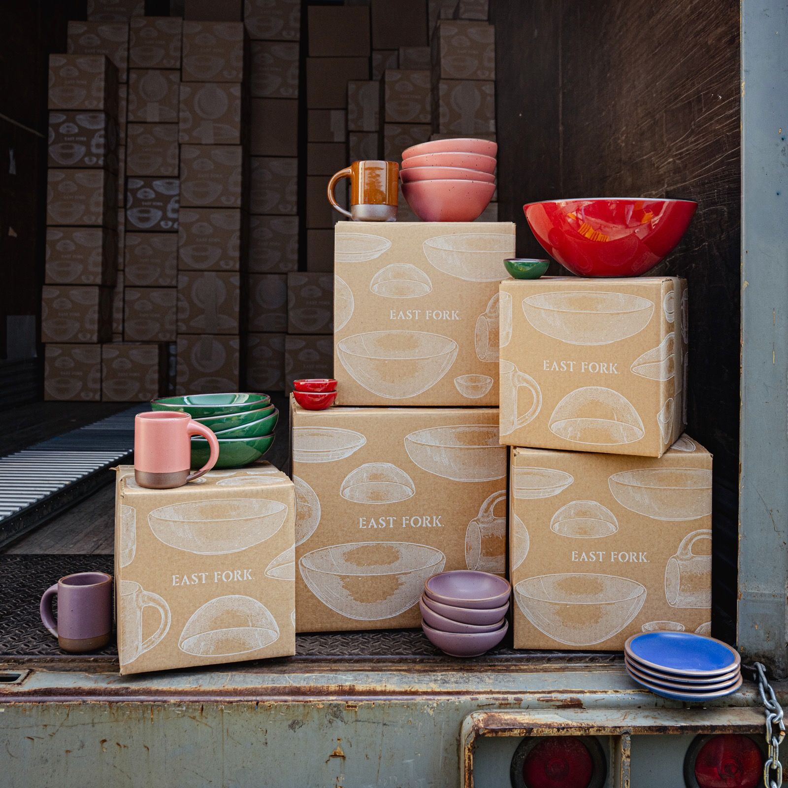 Stacks of East Fork shipping boxes topped with colorful ceramic bowls, plates, and mugs arranged inside a delivery truck.