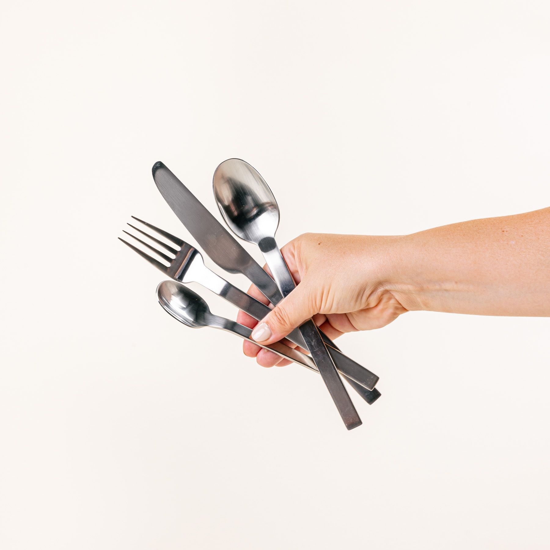 A hand holds a simple steel knife, fork, spoon, and teaspoon.