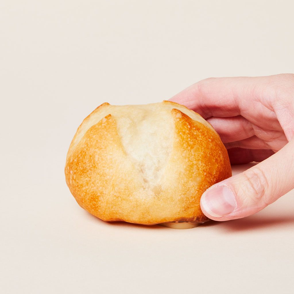 A hand grasps a light brown round object that has the markings and texture of a dinner roll