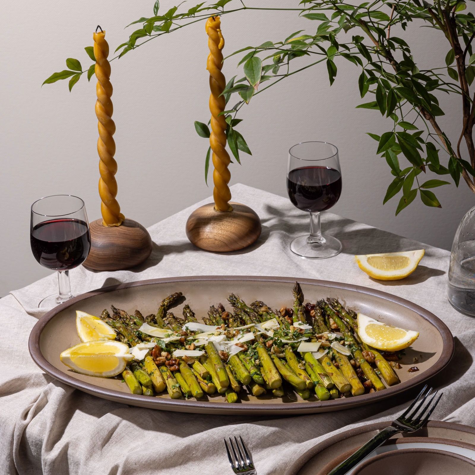 A ceramic oval platter in a taupe colors sits on a table with a spread of cooked asparagus and lemon slices. Surrounding is wine glasses, silverware, and twisted taper candles.
