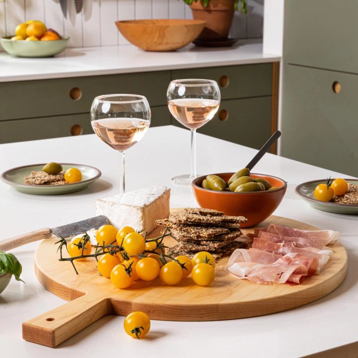 On a kitchen island is a round wooden serving board filled with charcuterie and surrounded by small plate, bowls, and wine glasses