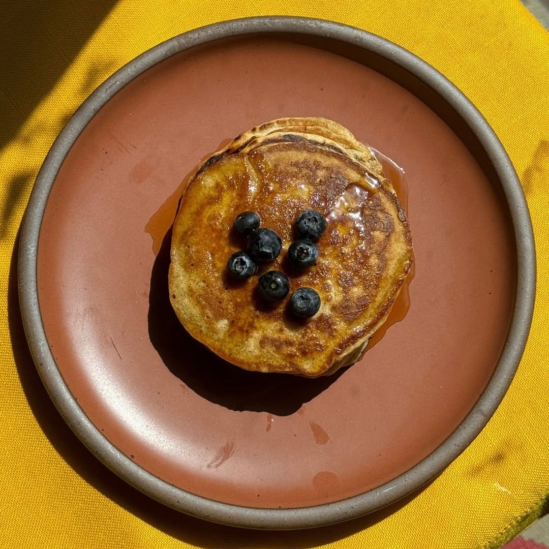 A medium cool terracotta ceramic plate with unglazed rim, plated with a stack of mini pancakes and blueberries.