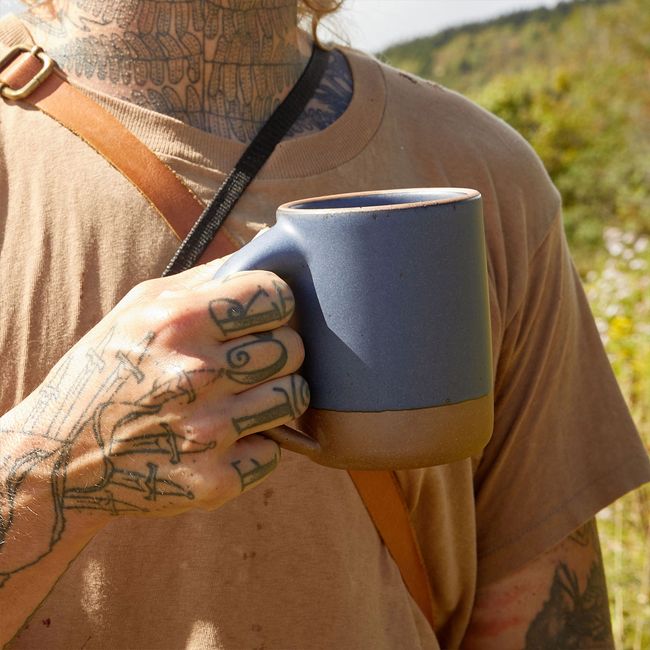 A tattooed hand  holds up an East Fork Mug in Blue Ridge, a muted navy color, in an outdoor setting with a grassy hill behind them.