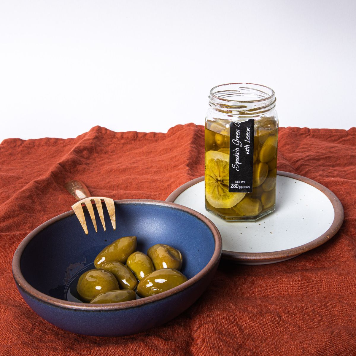 An open jar of olives with a blue bowl, white plate, brass fork - all sitting on a rust napkin