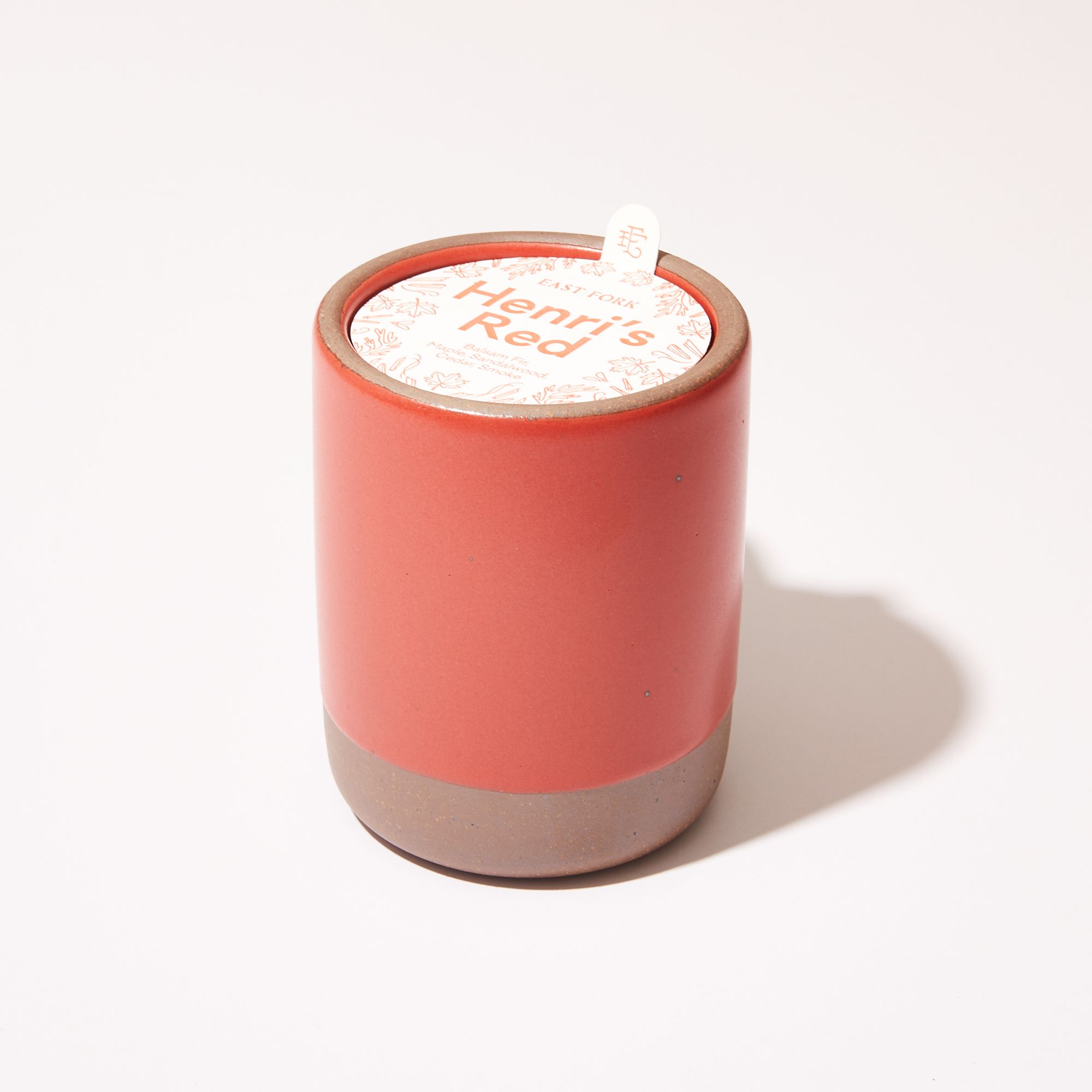 Large ceramic vessel in orange-red color with candle inside. On top is a packaging label sitting on top that reads "Henri's Red"