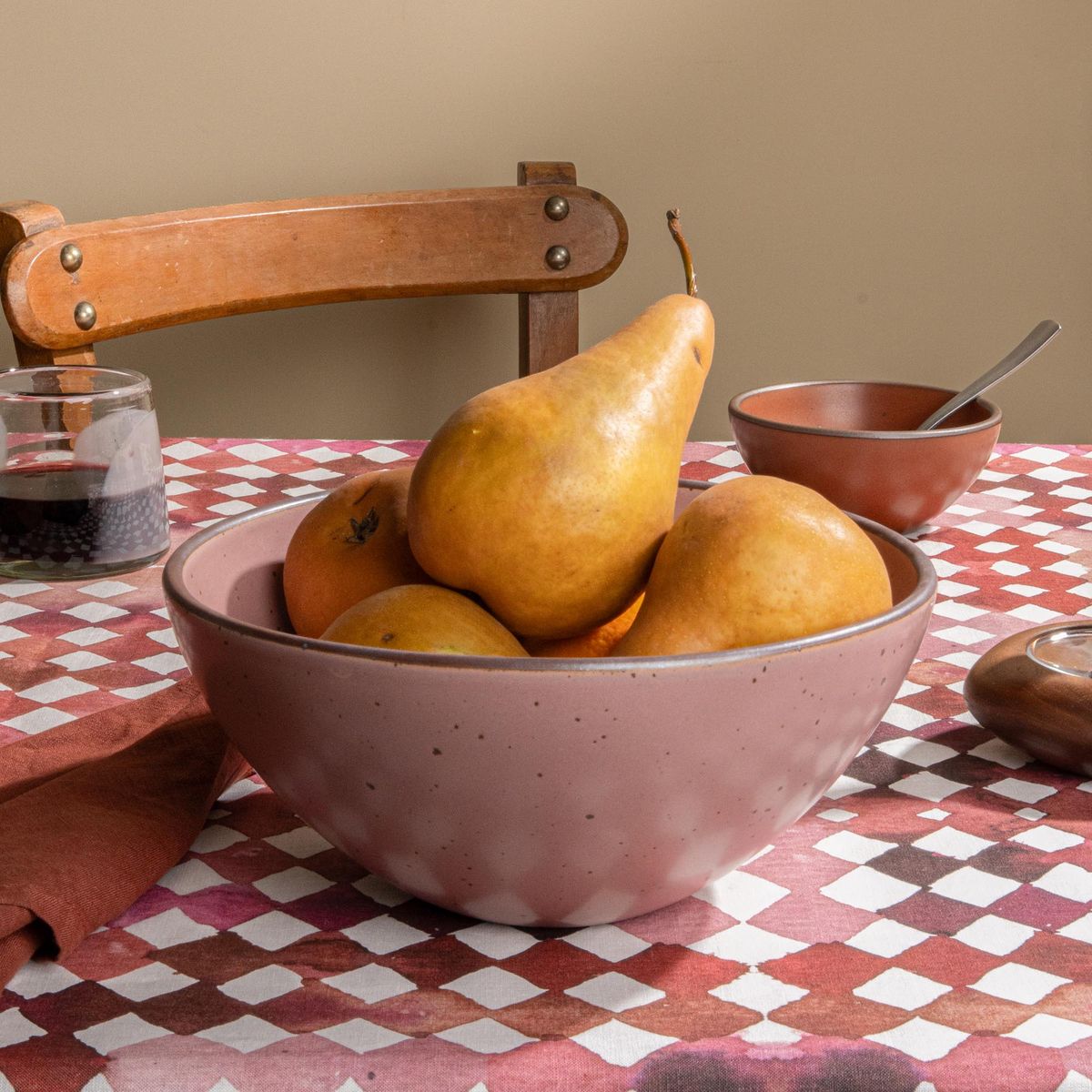 Dusty mauve pink ceramic fruit bowl filled with pears and citrus on a patterned tablecloth with a wooden chair in the background.